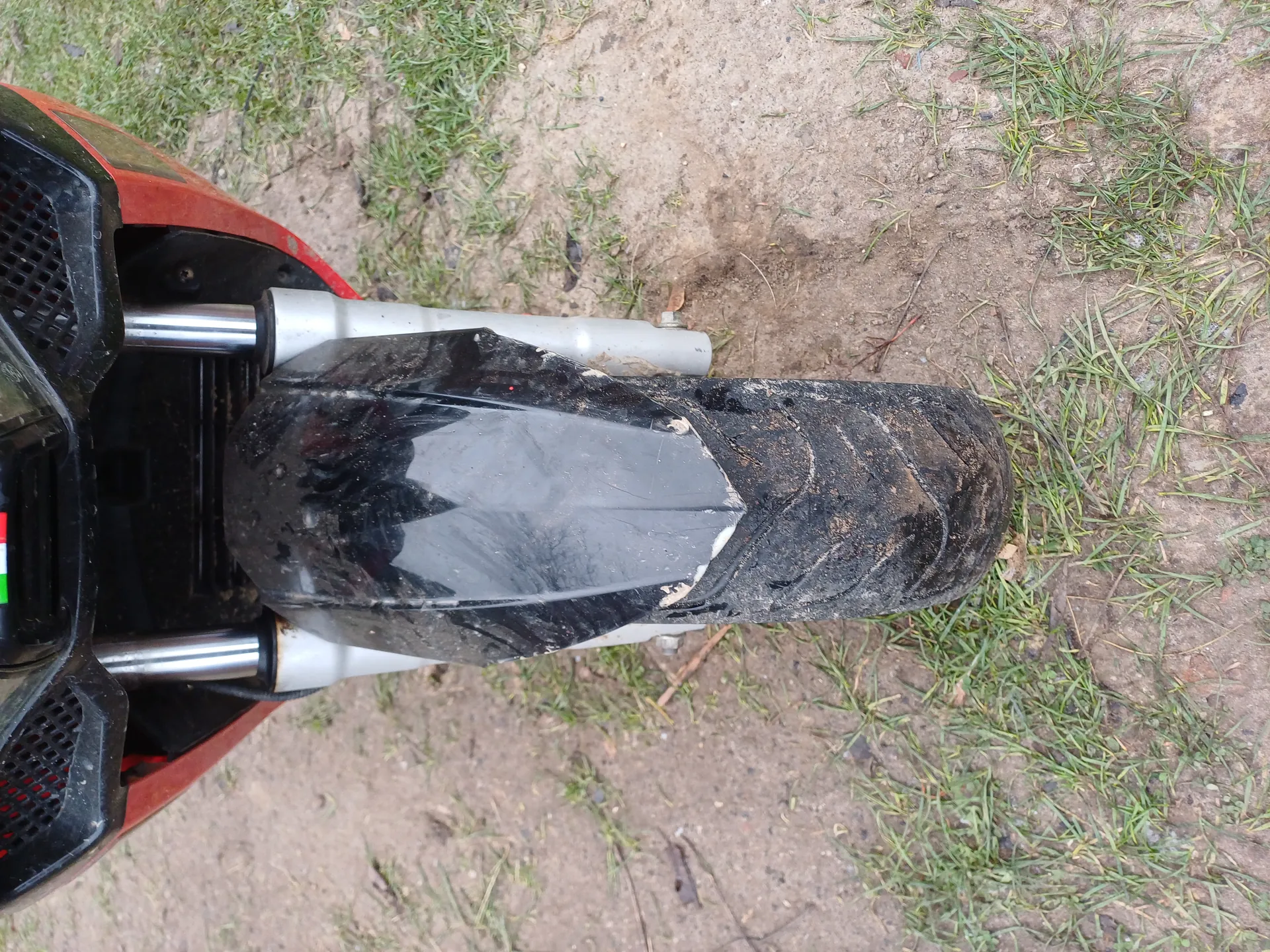 Zdjęcie przedstawia zbliżenie przodu motocykla z czarnym, zabrudzonym kołem przednim i częścią czerwonego nadwozia. Widoczne są srebrne widełki, czarna osłona oraz fragment ziemi z trawą i błotem. Na lewej stronie znajduje się mały naklejony proporczyk Włoch.