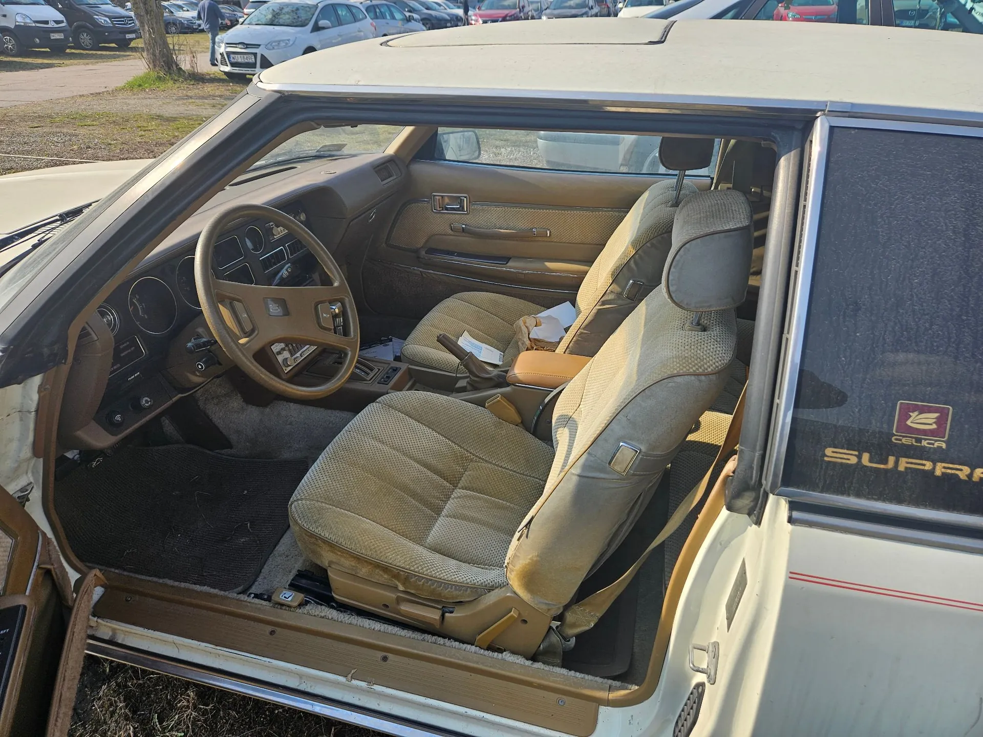 Wnętrze starego samochodu Toyota Celica Supra widziane z otwartych drzwi pasażera. Widoczna jest beżowa tapicerka foteli, kierownica, deska rozdzielcza oraz fragmenty nadwozia w kolorze białym. Na słupku przy drzwiach znajduje się logo Celica oraz napis SUPRA.