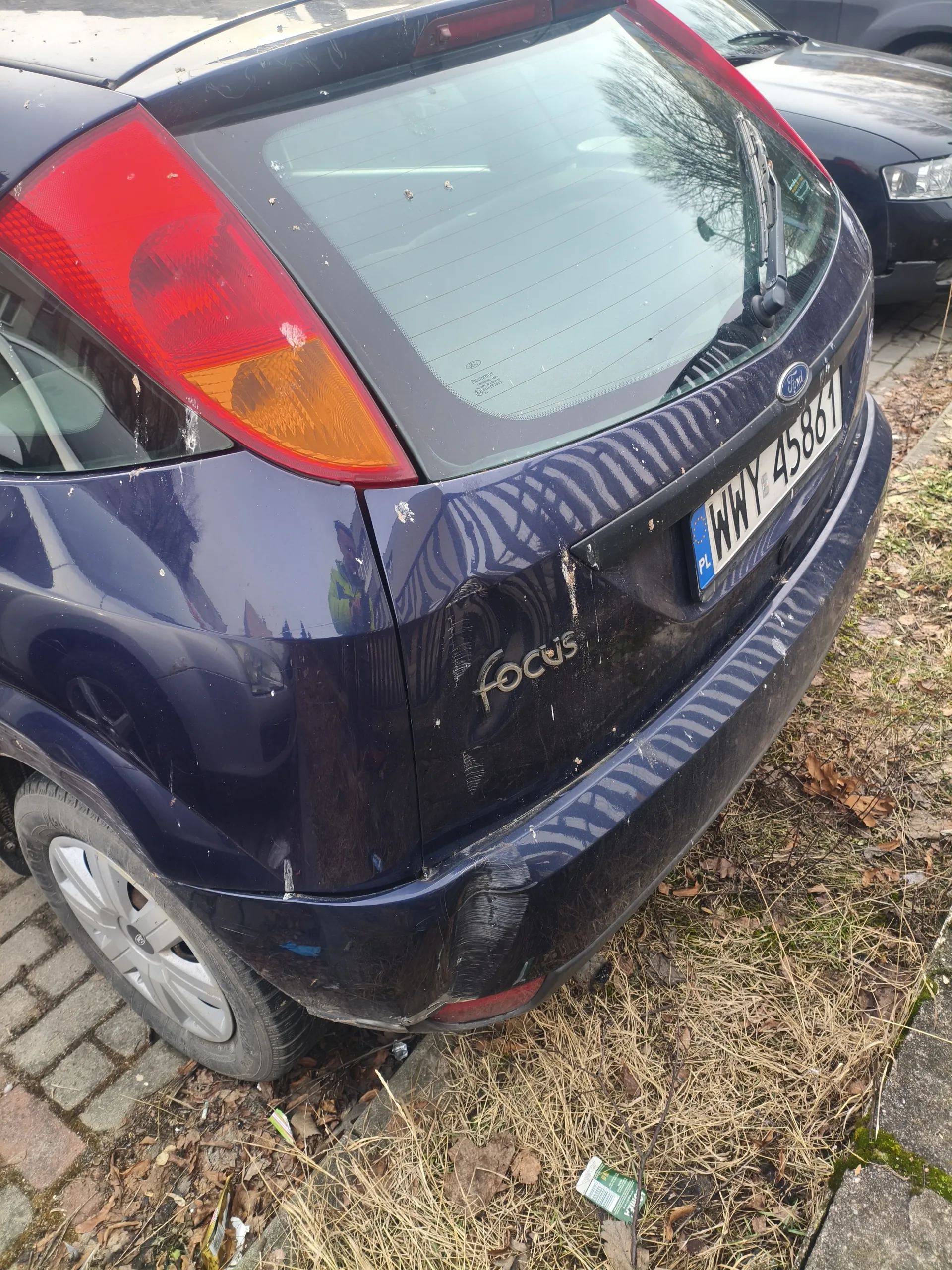 Z tyłu widoczny ciemnoniebieski samochód Ford Focus z widocznym napisem "focus" na bagażniku. Tablica rejestracyjna z białym tłem i czarnym tekstem "WWY 45861", z niebieskim pasem po lewej stronie z napisem "PL". Samochód ma zadrapania i zabrudzenia na karoserii. W tle częściowo widoczny inny czarny samochód. Podwozie stoi na brukowanej powierzchni obok suchego trawnika z rozsypanymi liśćmi i śmieciem. Czerwone światło tylnego reflektora i pomarańczowy kierunkowskaz są widoczne po lewej stronie.
