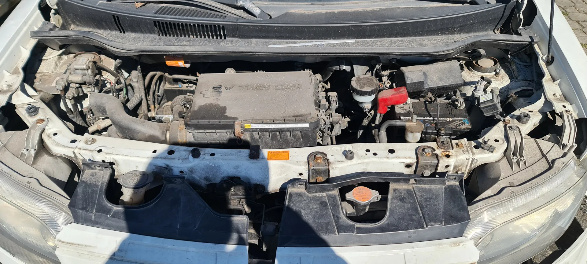 Otwarty kapota samochodu z widocznym silnikiem. Na pokrywie silnika widoczny napis '16V TWIN CAM'. W silniku znajdują się różne elementy, w tym czerwony korek, przewody, akumulator oraz reflektory po obu stronach. Widoczne są również ślady rdzy na metalowych elementach.