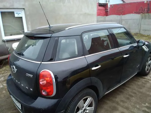 Czarny samochód Mini Cooper Countryman widziany od tyłu i boku. Na tylnej części widoczny napis 'COOPER' oraz logo marki. Samochód zaparkowany na podwórku z ziemią i żwirem. W tle betonowa ściana, okno z nieczytelnym napisem oraz czerwona budowla. Tablica rejestracyjna na tylnym zderzaku jest częściowo widoczna.