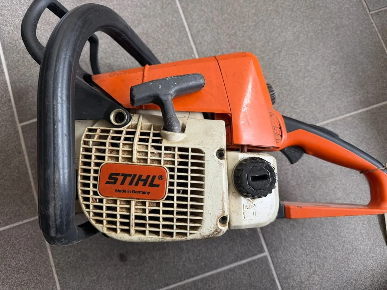Pomarańczowo-biała piła spalinowa marki Stihl leżąca na szarych płytkach. Na białej obudowie silnika znajduje się logo 'STIHL' z dopiskiem 'Made in Germany'. Urządzenie posiada czarne uchwyty i czarną zakrętkę zbiornika paliwa.
