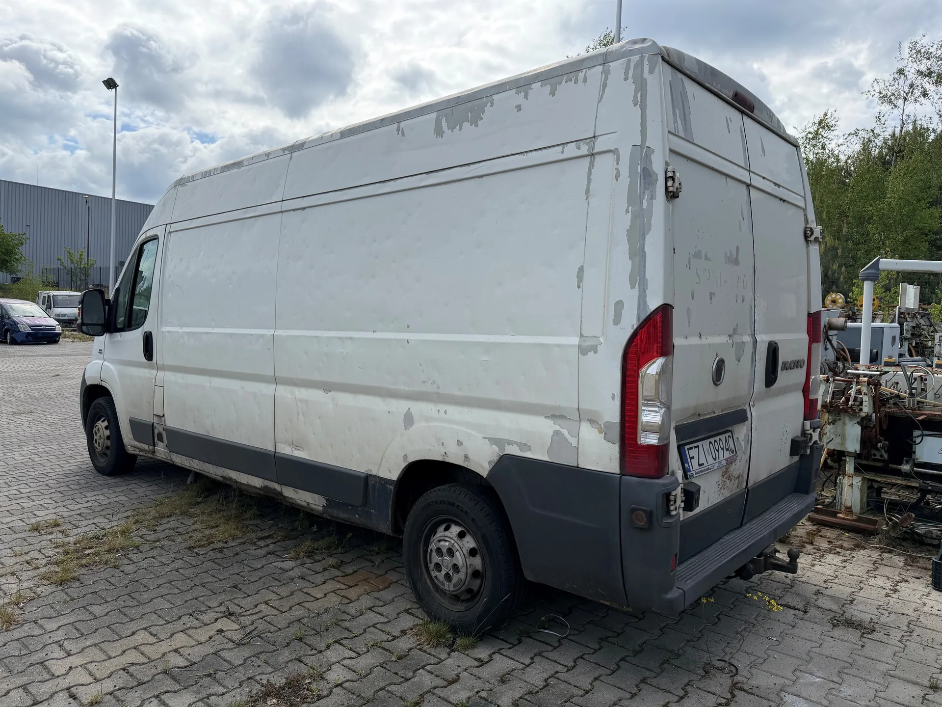 Biały dostawczy van z widocznymi uszkodzeniami i obłupioną farbą, zaparkowany na brukowanej powierzchni z trawą między kamieniami. Na tylnym zderzaku tablica rejestracyjna FZ 099 AC. W tle budynki przemysłowe, inne pojazdy (m.in. niebieski samochód osobowy), maszyny przemysłowe oraz drzewa. Niebo częściowo zachmurzone. Van ma szare elementy na dole i czerwone światła tylnego oświetlenia.