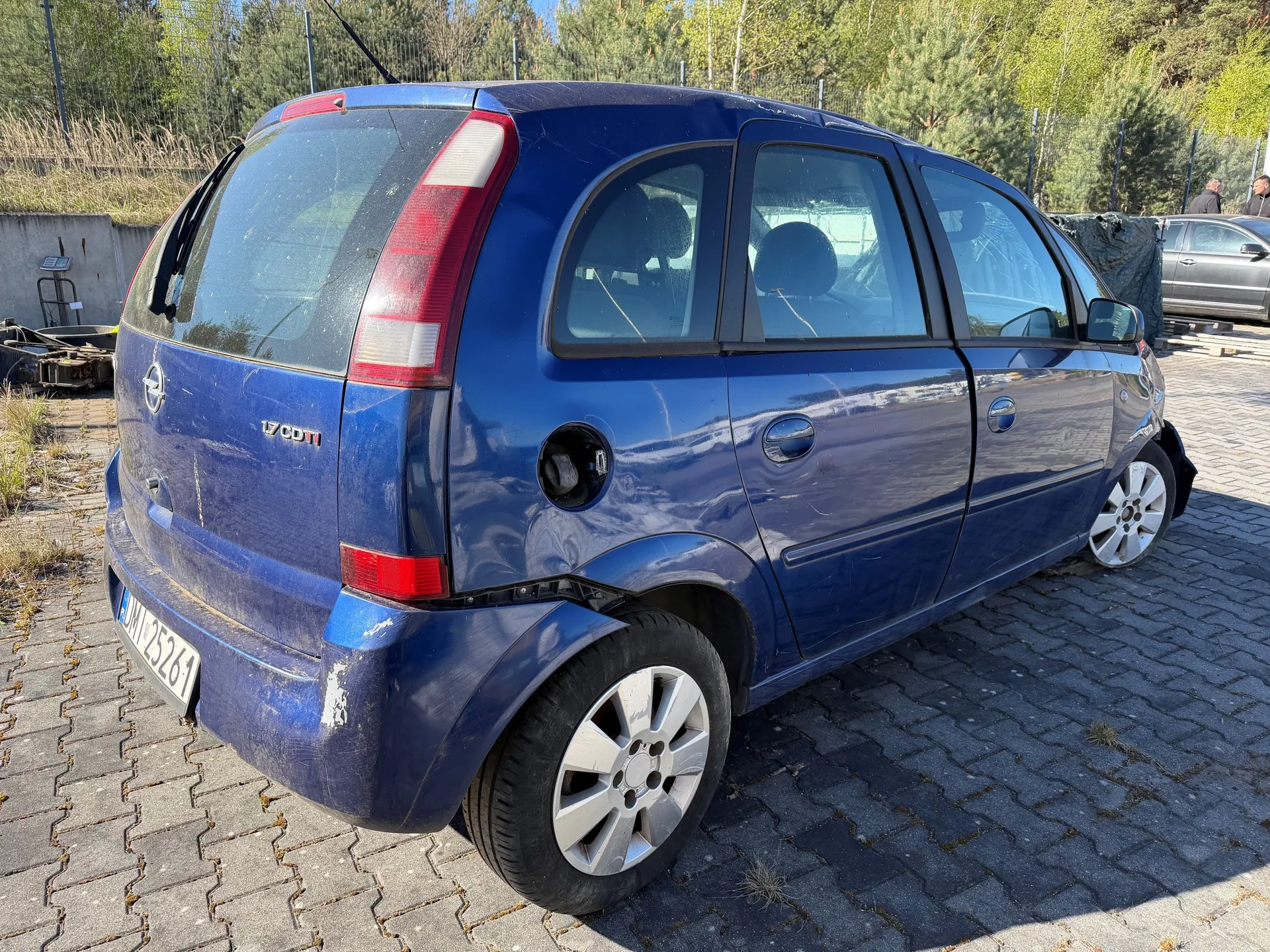 Niebieski samochód osobowy typu hatchback z widocznym tyłem i bokiem. Na tylnej szybie napis "1.7 CDTI", tablica rejestracyjna "DM 25261". Samochód ma srebrne felgi, czerwone światła tylnego i widoczne ślady zabrudzeń na karoserii. W tle: brukowana powierzchnia, metalowe ogrodzenie, drzewa, częściowo zakryty inny samochód oraz dwie osoby stojące w oddali.