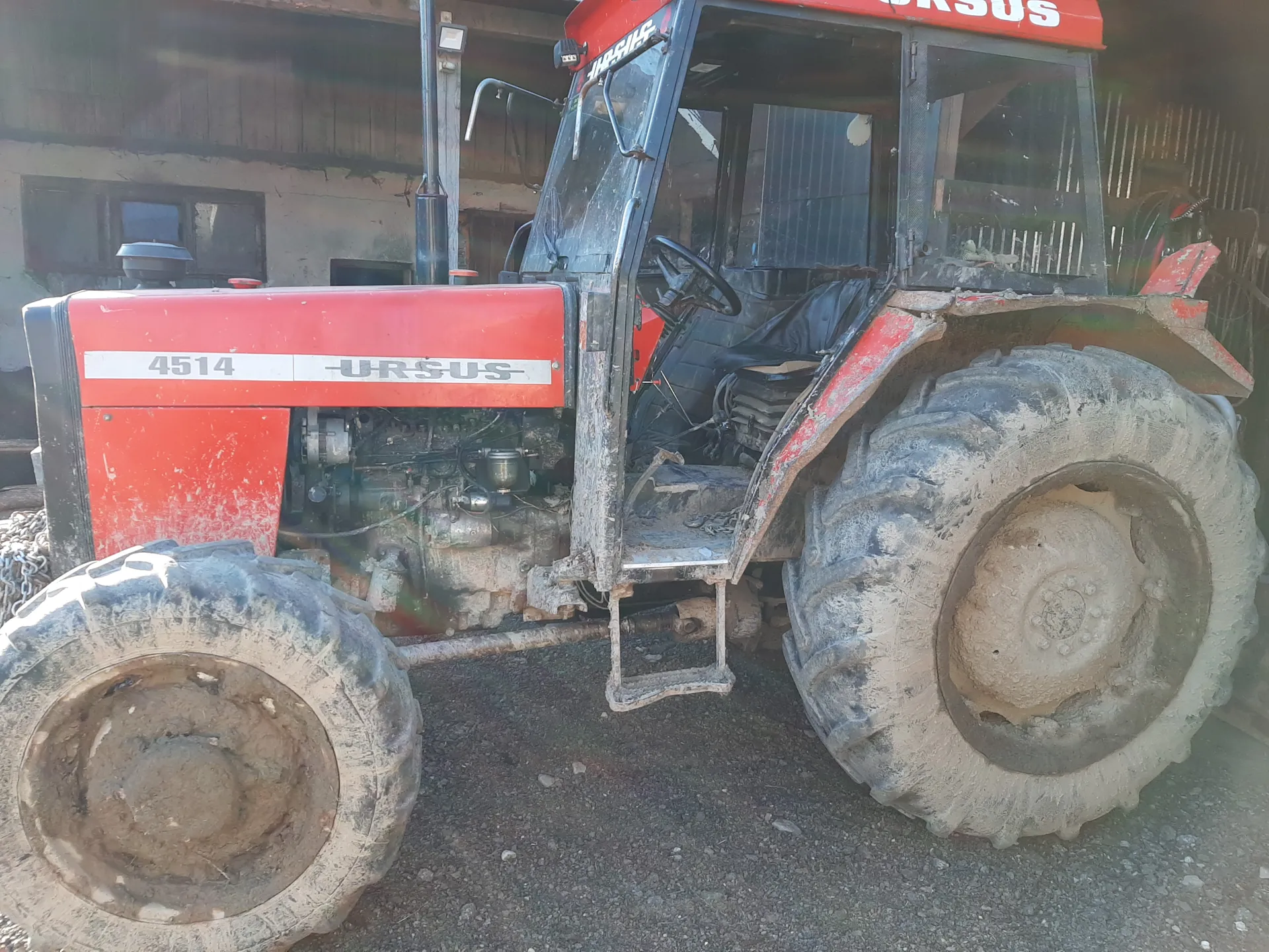 Czerwony traktor marki URSUS model 4514 z widocznym numerem i nazwą na boku. Traktor pokryty jest błotem, szczególnie na dużych oponach. W tle stara, drewniana budowla z oknami. Wnętrze kabiny z kierownicą i czarnym siedzeniem jest częściowo widoczne. Traktor stoi na podłożu z drobnych kamieni.
