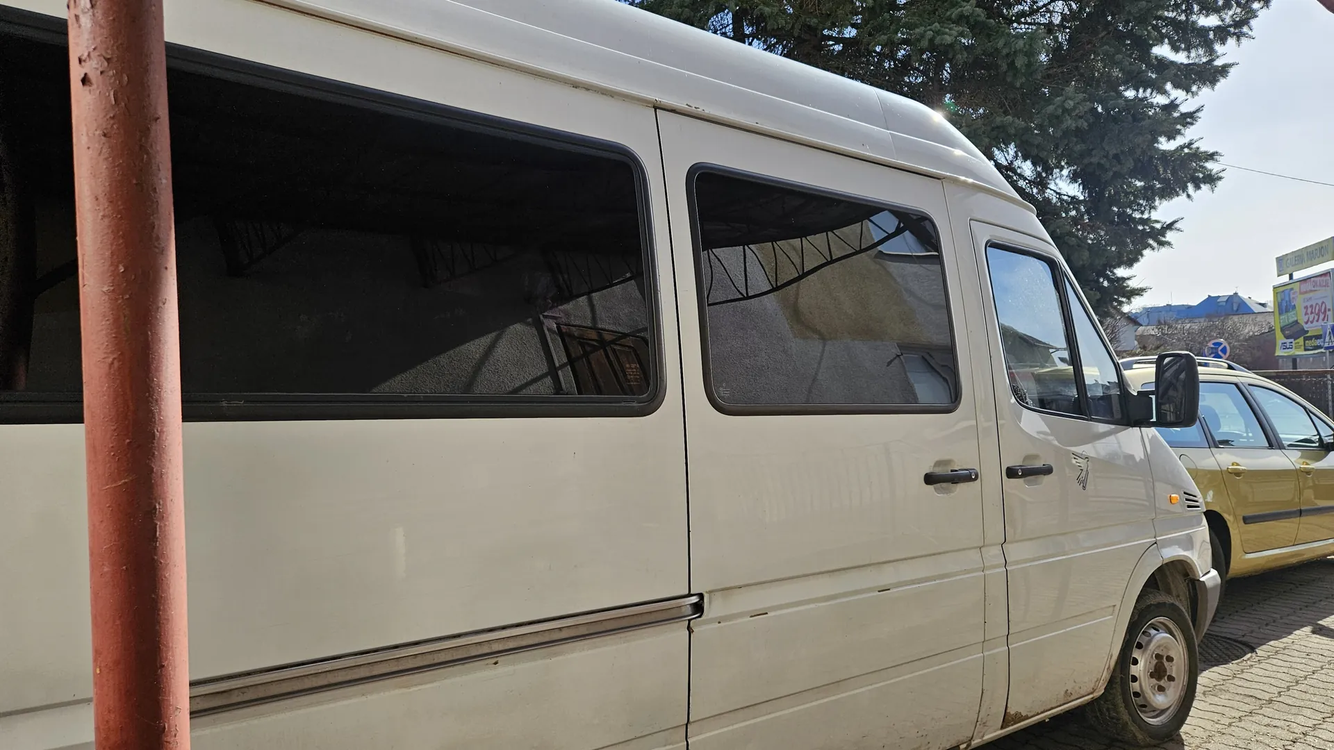 Biały furgon zaparkowany na brukowanej ulicy z czerwoną słupem po lewej stronie. Widoczne przyciemniane okna z odbiciami budynków i konstrukcji metalowych. Obok furgona żółty samochód osobowy. W tle drzewa, niebieski budynek oraz tablica reklamowa z napisem "3399". Niebo jest jasne.