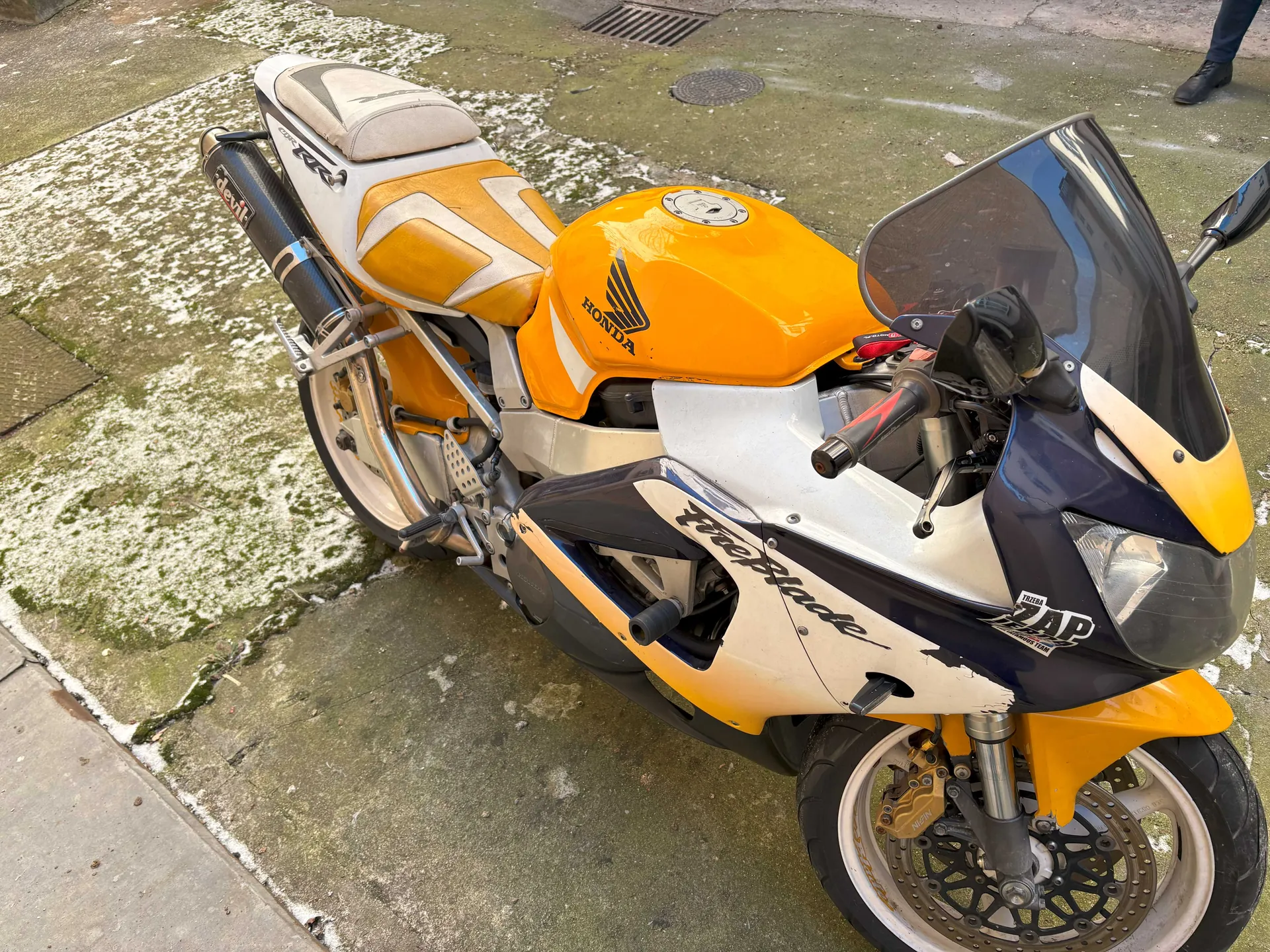 Motocykl marki Honda Fireblade w kolorach pomarańczowym, białym i ciemnoniebieskim, zaparkowany na betonowej powierzchni z plamami śniegu. Widoczne elementy: logo Honda na zbiorniku paliwa, napis Fireblade na boku, naklejka ZAP na przedniej części, wydech z napisem Akrapovič, reflektor, szyba przednia oraz fragment nogi osoby w czarnych butach w prawym górnym rogu. Tło: betonowa nawierzchnia z kanałem i pokrywką studzienki.