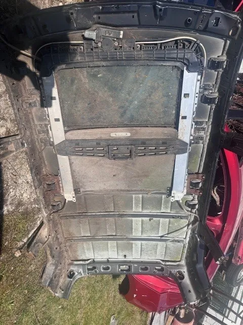 Zdjęcie przedstawia metalową konstrukcję, prawdopodobnie tylną część drzwi lub bagażnika samochodu, z widocznymi panelami, rdzą i przewodami elektrycznymi. W tle czerwony samochód, trawa i metalowe ogrodzenie. Brak czytelnego tekstu.