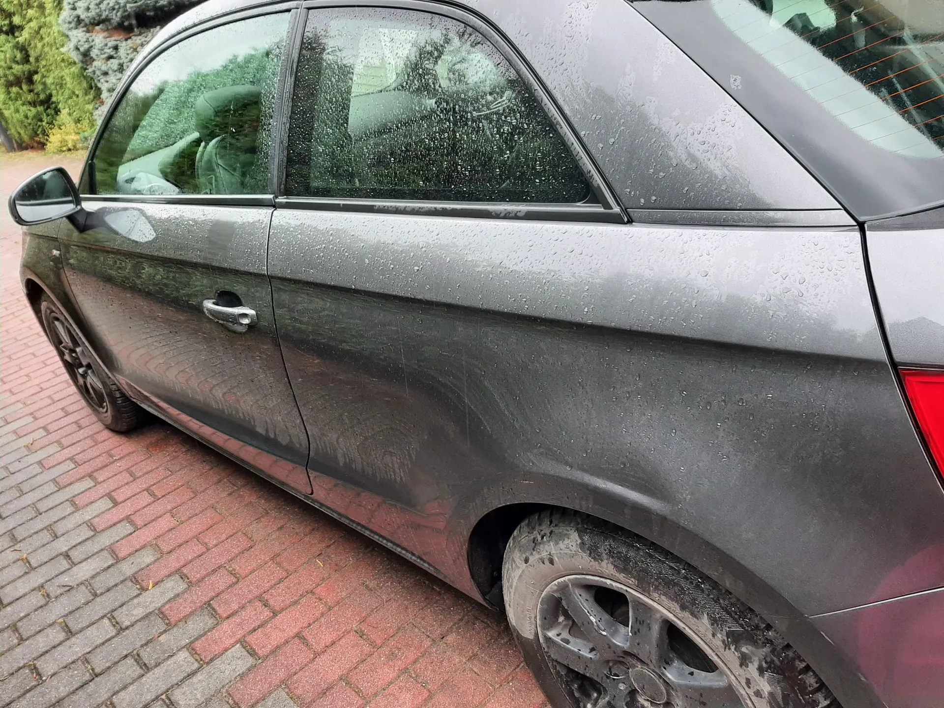 Zbliżenie na bok ciemnoszarego samochodu zaparkowanego na kostce brukowej. Karoseria auta jest pokryta kroplami wody. Widoczne są drzwi kierowcy, klamka, tylne koło z ciemną felgą oraz fragment tylnego światła. W tle widać zielone krzewy.