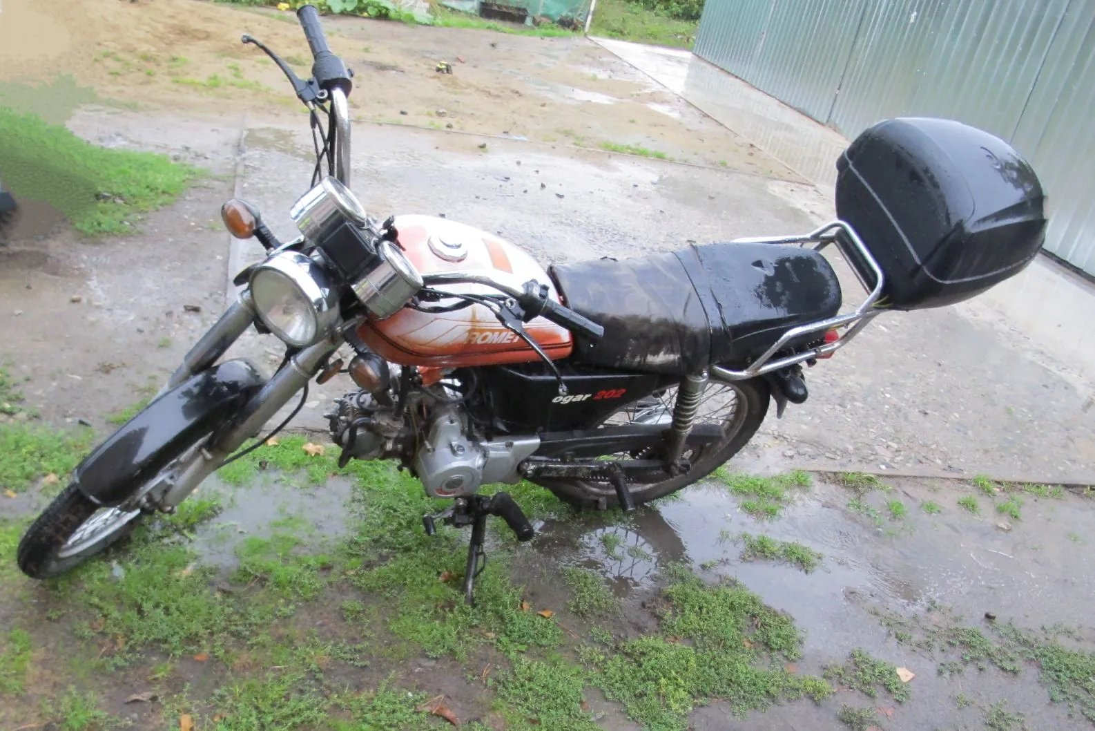 Pomarańczowo-czarny motocykl marki Romet Ogar 200 zaparkowany na mokrym, betonowym podjeździe z kępami trawy. Motocykl posiada czarne siedzenie, czarny kufer zamontowany z tyłu oraz okrągły reflektor z przodu. Na baku widnieje napis 'ROMET', a na bocznej osłonie 'ogar 200'.