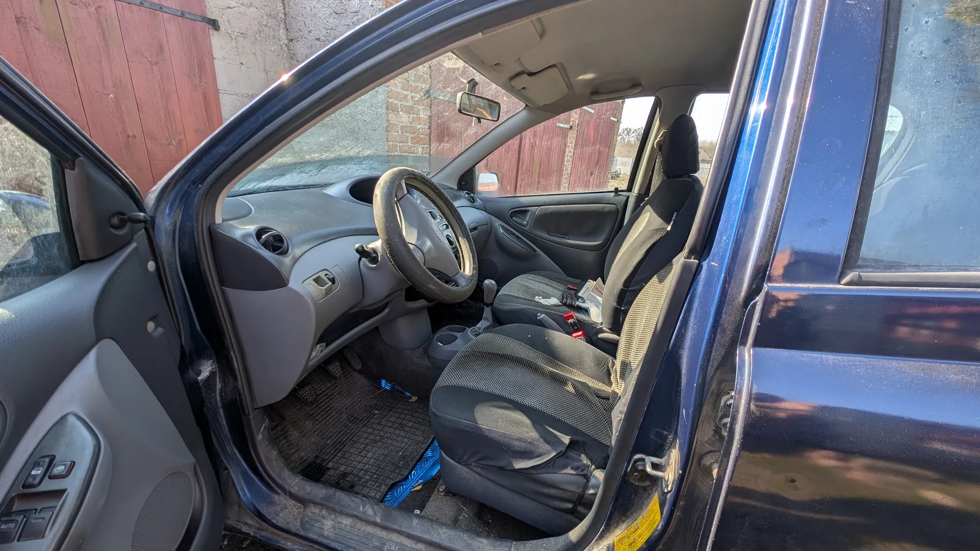Wnętrze niebieskiego samochodu z otwartą przodem drzwiami kierowcy. Widoczne są kierownica, deska rozdzielcza, czarne siedzenia z siateczkowym wzorem, podłoga z czarnym dywanikiem i niebieskim przedmiotem. Na panelu drzwiowym znajdują się przyciski sterujące. W tle ceglana ściana i czerwone drewniane drzwi garażowe. Samochód znajduje się na zewnątrz, w słoneczny dzień.