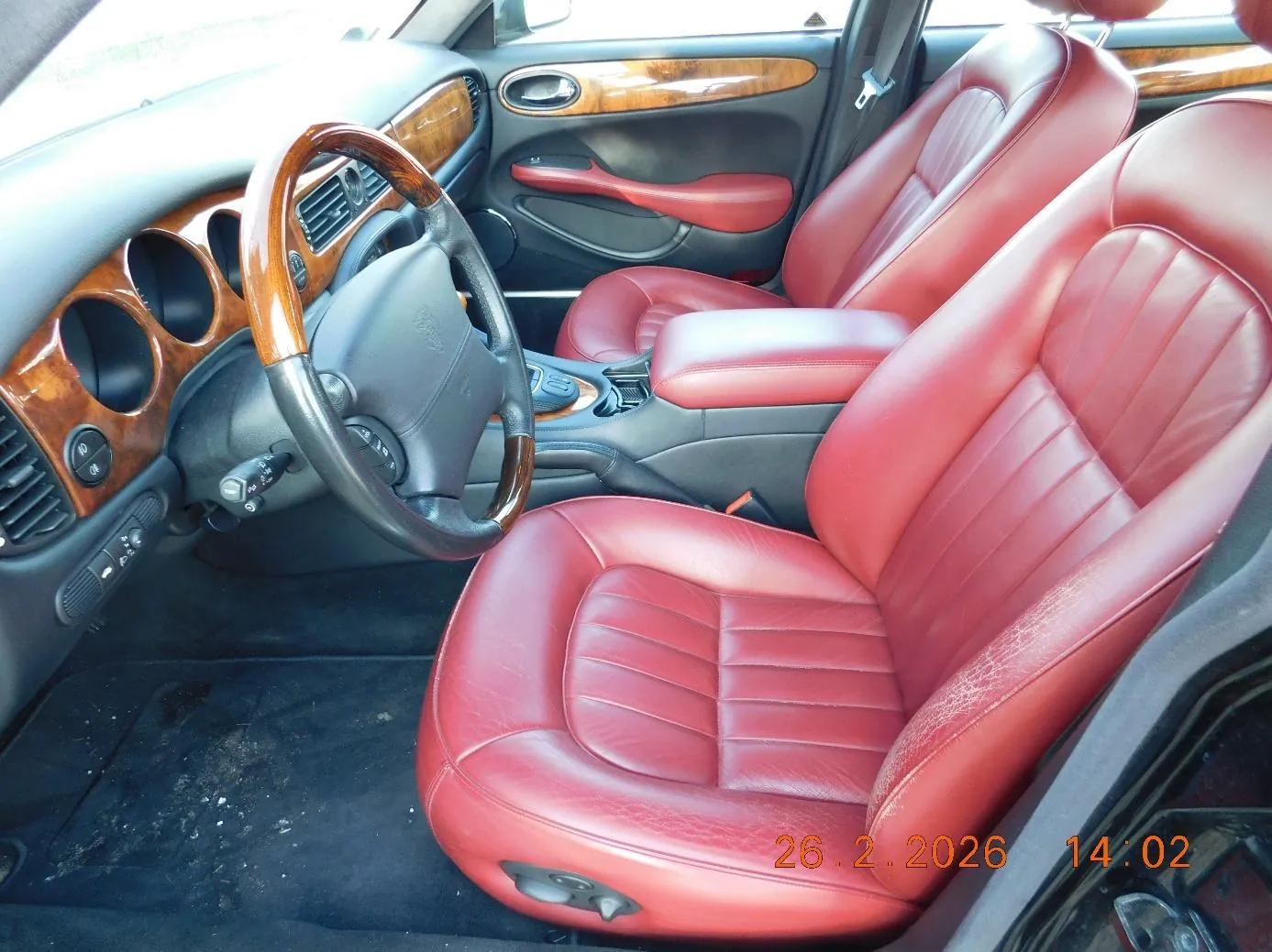Wnętrze samochodu z czerwonymi skórzany siedzeniami, kierownicą z drewnianym obwodem, deska rozdzielcza z drewnianymi wstawkami i okrągłymi otworami wentylacyjnymi. Widoczne są drzwi z czerwonymi podłokietnikami i drewnianymi elementami. Na dole obrazu znajduje się pomarańczowy znaczek z datą 26.2.2026 i godziną 14:02.