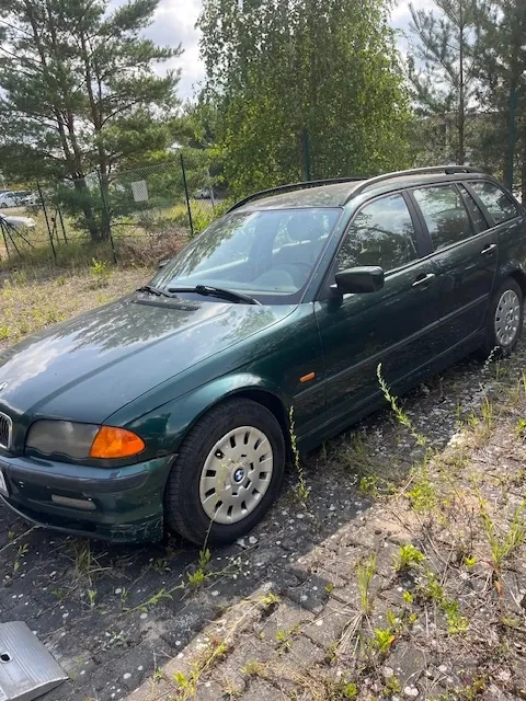 Ciemnozielony samochód osobowy typu kombi marki BMW zaparkowany na zniszczonym podjeździe z kostki brukowej, z której wyrasta trawa. Auto jest widoczne z perspektywy przedniego lewego boku. W tle znajdują się zielone drzewa i ogrodzenie z siatki.