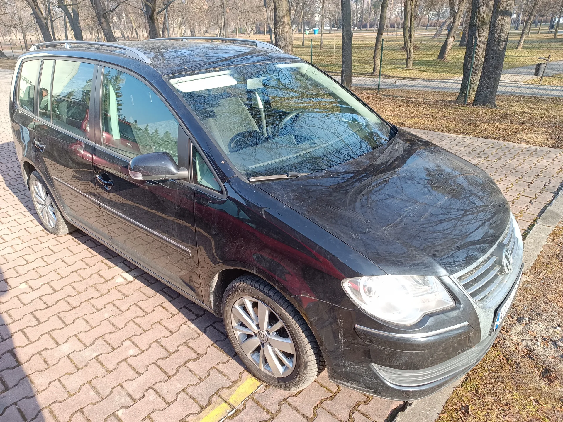 Czarny samochód osobowy typu minivan marki Volkswagen zaparkowany na brukowanej powierzchni. W tle drzewa bez liści, siatka ogrodzeniowa i trawiaste pole. Samochód posiada dachowy bagażnik, felgi aluminiowe oraz reflektory. Słoneczny dzień, widoczne cienie na ziemi i odbicia na karoserii.