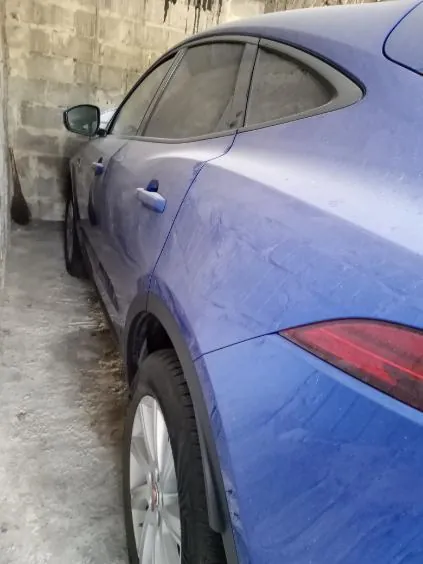 Niebieski samochód osobowy widziany z boku i tyłu, zaparkowany przy betonowej ścianie z bloczków. Samochód ma widoczne zadrapania na karoserii, czerwone światło tylnego reflektora oraz srebrne felgi. Tło stanowi betonowa ściana z ciemnymi plamami i betonowa podłoga.