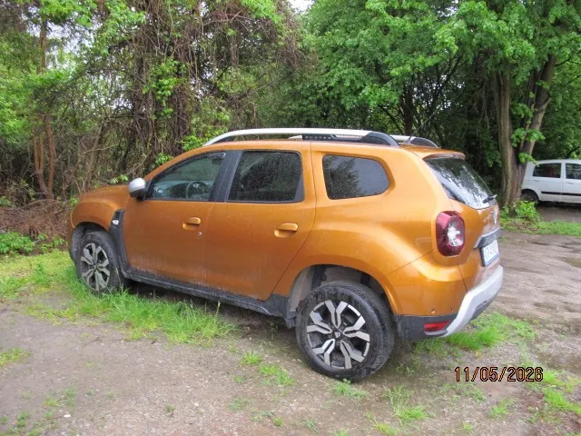 Pomarańczowy SUV z białym dachem i aluminiowymi felgami zaparkowany na żwirowym obszarze z trawą. W tle zielone drzewa i biały samochód. W prawym dolnym rogu widoczna data 11/06/2026.