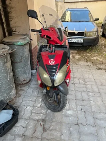Czerwono-czarny skuter z logo na przodzie zaparkowany na brukowanej powierzchni. Za skutrem widoczny ciemny samochód osobowy marki Skoda z tablicą rejestracyjną WPU 3487. Po lewej stronie obrazu dwa duże metalowe pojemniki na śmieci, jeden z zielonym kapturem. Tło stanowi fragment budynku i trawnik.