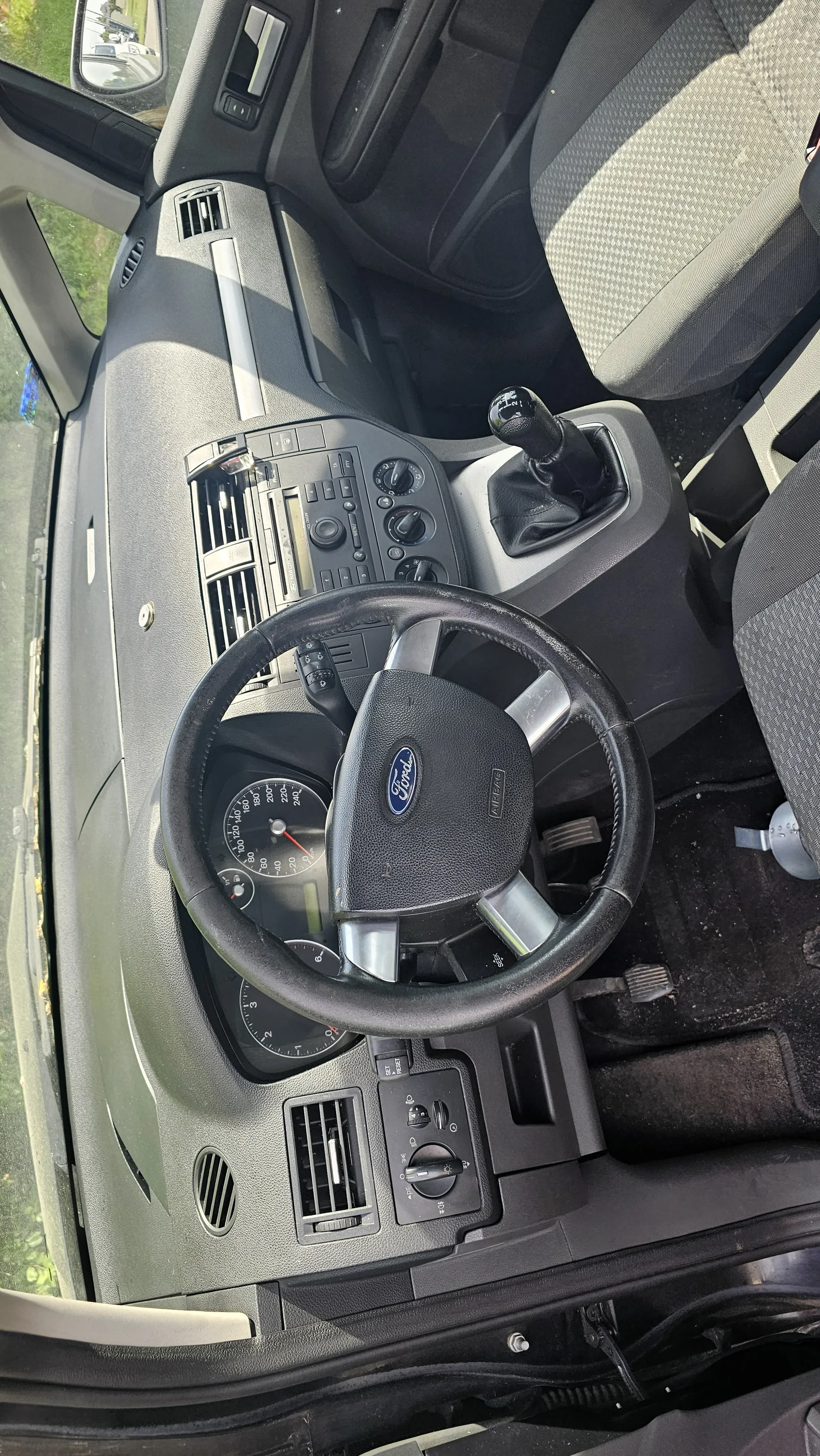 Wnętrze samochodu widziane z góry. Kierownica z logo Ford w centrum, deska rozdzielcza z prędkościomierzem, tachometrem, wentylacjami i systemem radiowym. Dźwignia zmiany biegów po lewej stronie kierownicy, siedzenia z tkaniną w wzór, pedały i dywanik na podłodze. Widoczne drzwi samochodu i lusterko boczne.