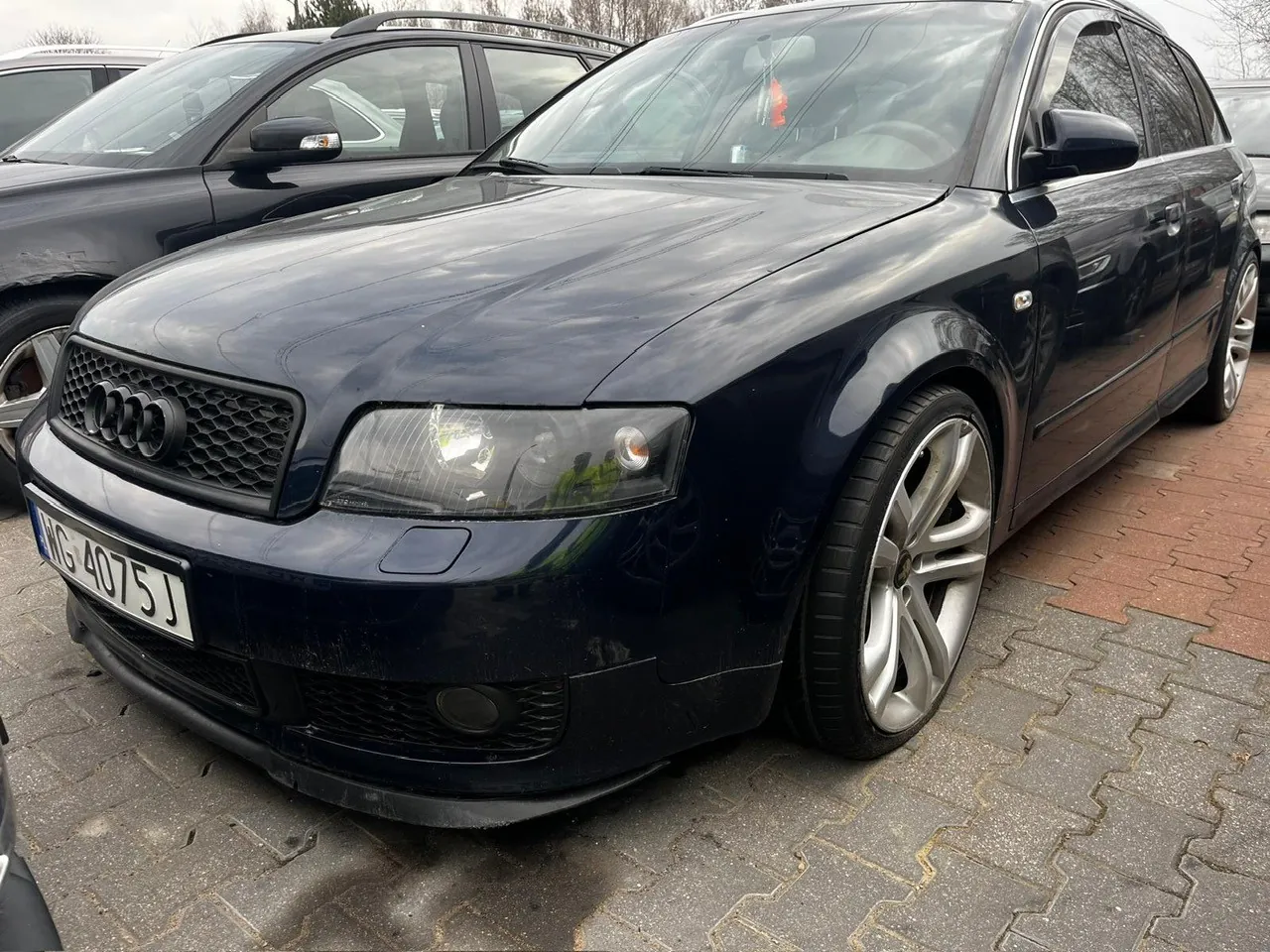 Ciemnoniebieski samochód Audi A6 Avant zaparkowany na brukowanej powierzchni. Widoczna tablica rejestracyjna z numerem WG 4075 J. W tle częściowo widoczny inny ciemny samochód oraz drzewa i chmurne niebo. Samochód ma srebrne felgi i czarną atrapę chłodnicy z logo Audi.