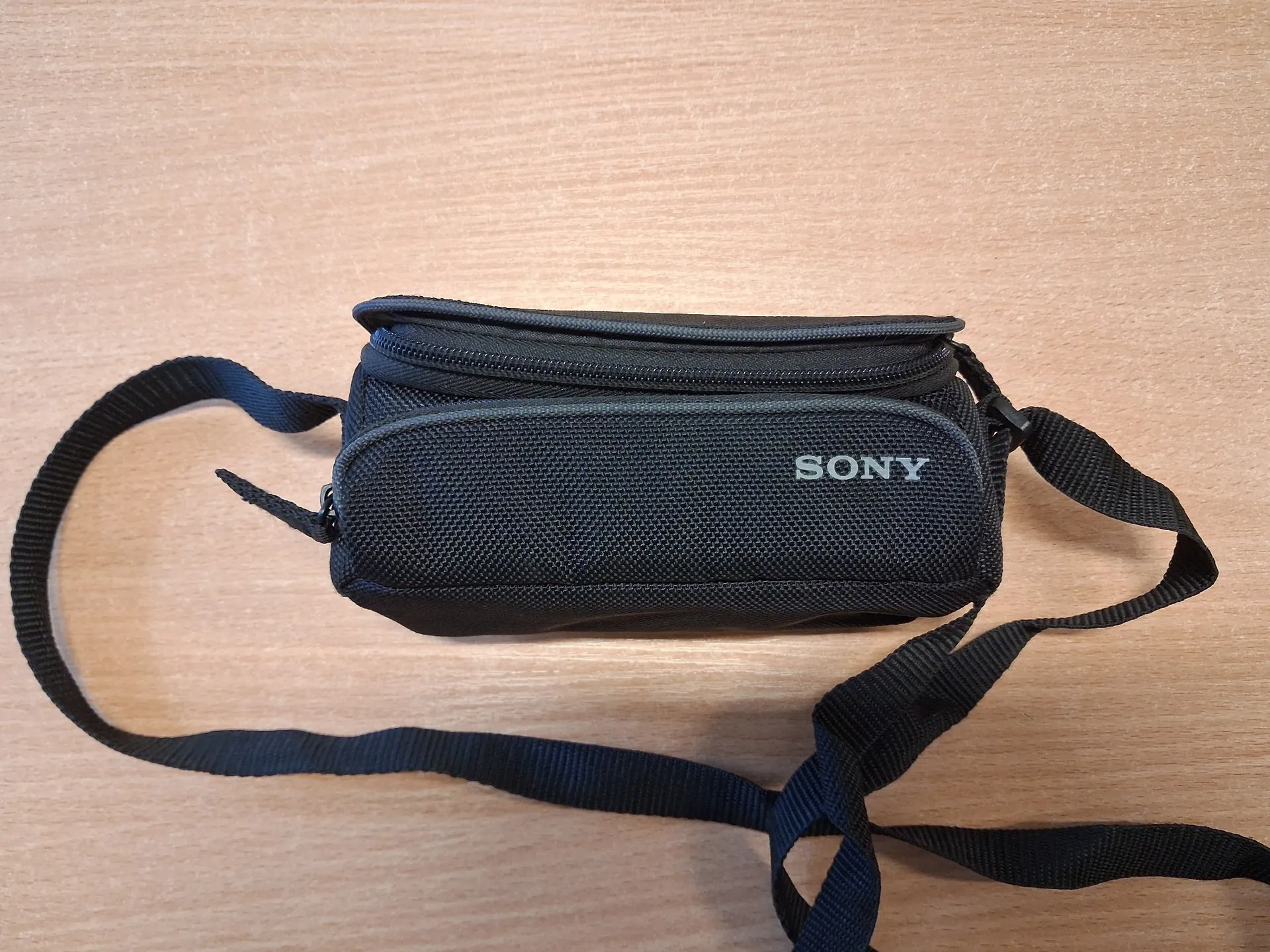 Czarna, materiałowa torba na aparat marki Sony z białym napisem 'SONY' na przedniej ściance. Torba posiada zamek błyskawiczny na górze i czarny pasek do przenoszenia, który jest rozłożony wokół niej. Całość znajduje się na jasnobrązowym, drewnianym blacie.