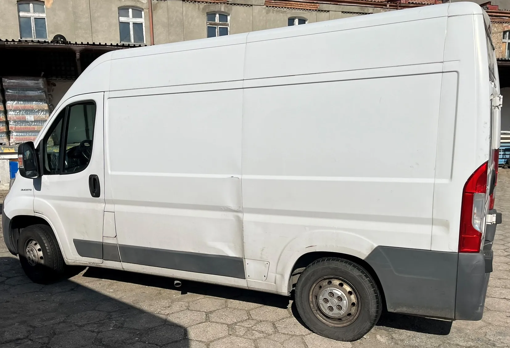 Biały furgon zaparkowany na kostce brukowej na tle budynku. Pojazd ma widoczne wgniecenie na dolnej części bocznego panelu nad tylnym kołem oraz wzdłuż środkowej części nadwozia.
