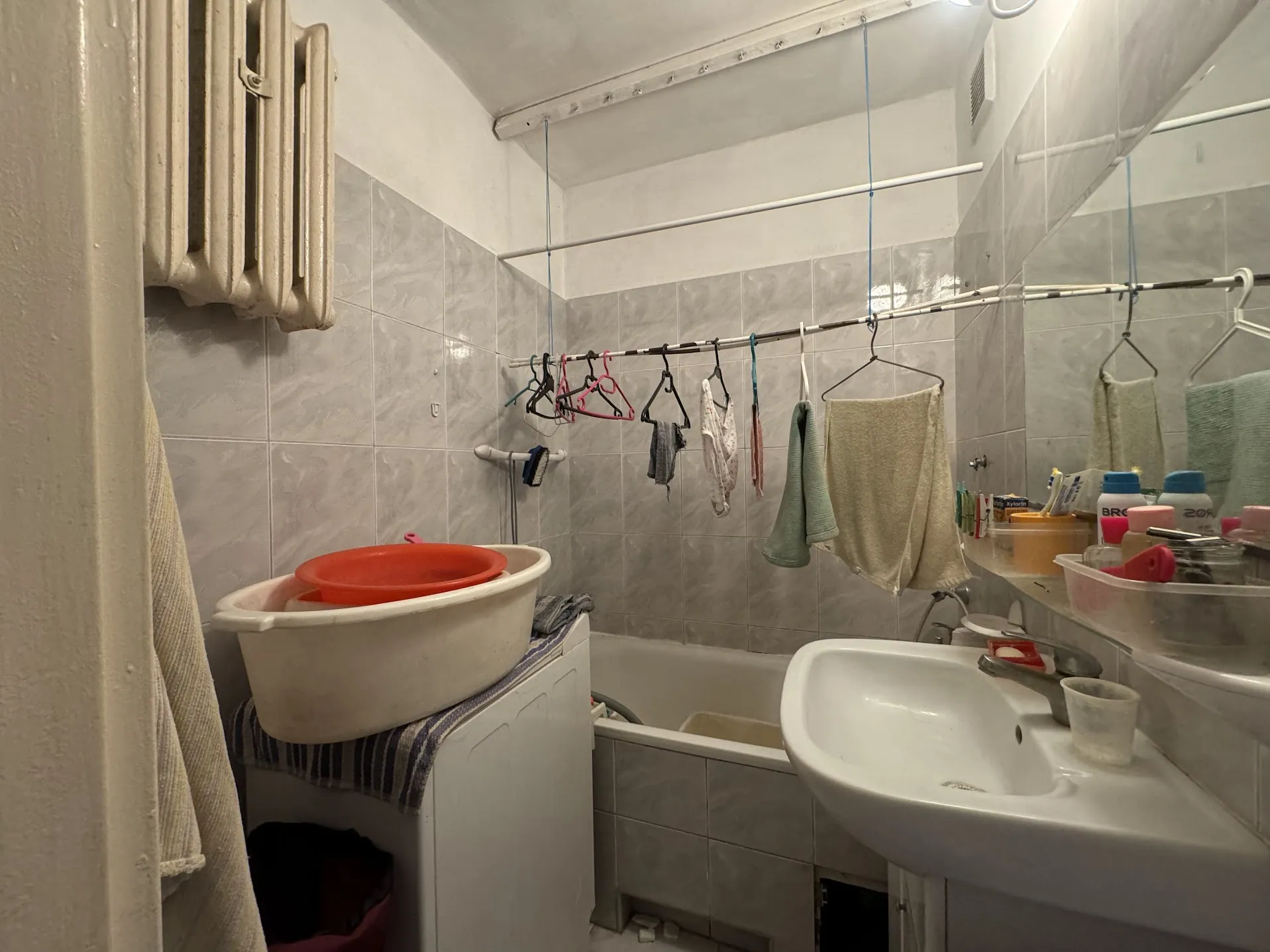 Wnętrze łazienki z białą umywalką, wanną i pralką. Na pralce znajdują się biała i czerwona miska. Na ścianie wiszą suszarka z różowymi i czarnymi wieszakami oraz ręczniki. Na półce obok umywalki widoczne butelki z napisami "BRO" i "20", pojemniki i inne artykuły higieniczne. Ściany wyłożone szarymi płytkami, a na lewej stronie znajduje się grzejnik. Oświetlenie sufitowe.