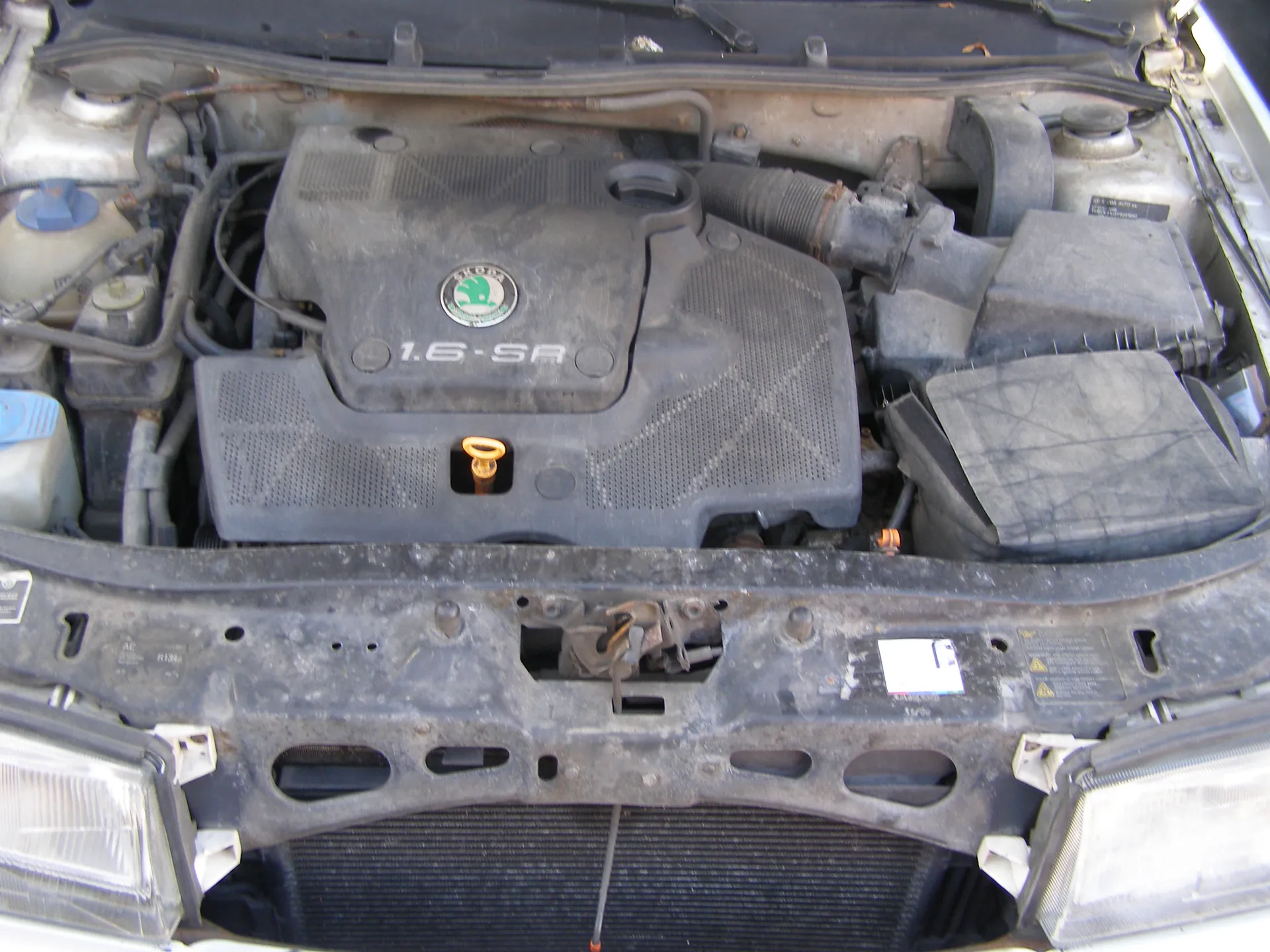Widok otwartej tylnej części silnika samochodu marki Skoda. Na pokrywie silnika widoczny jest zielony logo Skody oraz napis "1.6-SR". Silnik i otaczające go elementy są zabrudzone. Po lewej stronie znajduje się zbiornik chłodniczy z niebieskim korkiem, a po prawej czarna skrzynka. W dolnej części obrazu widoczne są reflektory oraz zardzewiały zderzak z naklejkami i ostrzeżeniami. W centrum znajduje się żółty korek do oleju.