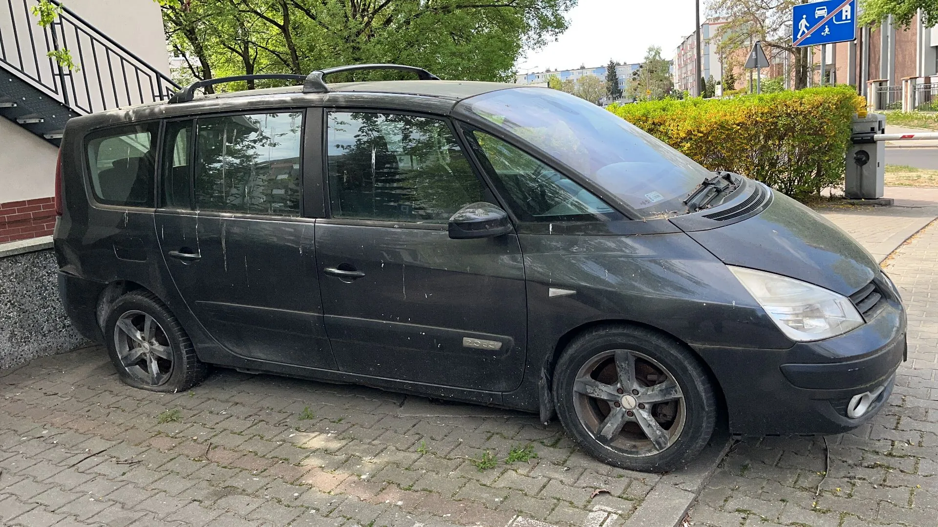 Czarny, zabrudzony samochód typu minivan zaparkowany na kostce brukowej. Pojazd posiada relingi dachowe i pięcioramienne felgi aluminiowe. W tle widoczne są zielone drzewa, krzewy oraz fragmenty budynków i znak drogowy informujący o przejściu dla pieszych.