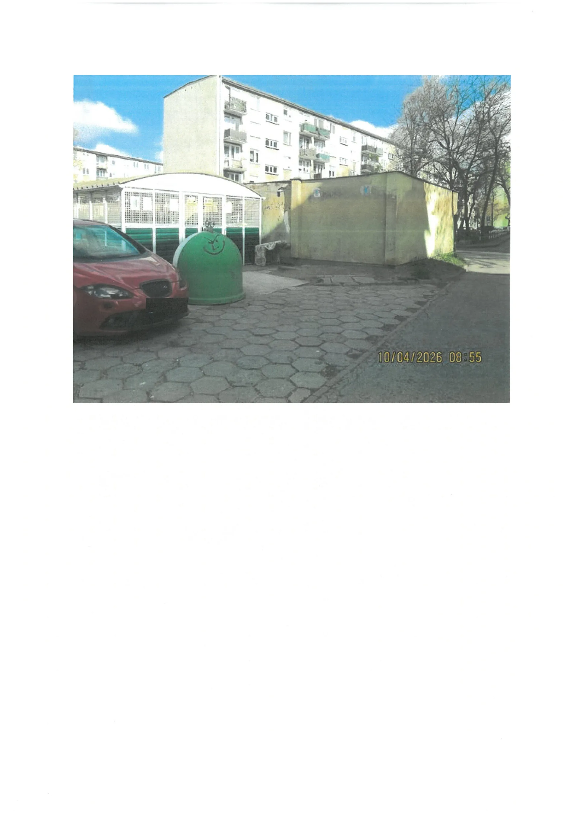 Zdjęcie przedstawia fragment osiedla mieszkaniowego. Na pierwszym planie po lewej stronie widoczny jest przód czerwonego samochodu zaparkowanego na placu wyłożonym szarą kostką brukową w kształcie sześciokątów. Obok samochodu stoi duży, zielony, kulisty pojemnik na odpady. W tle znajduje się niski, żółty budynek gospodarczy oraz wielopiętrowy blok mieszkalny z jasną elewacją. Po prawej stronie widoczne są drzewa bez liści. W prawym dolnym rogu zdjęcia znajduje się żółty napis z datą i godziną: 10/04/2026 08:55.