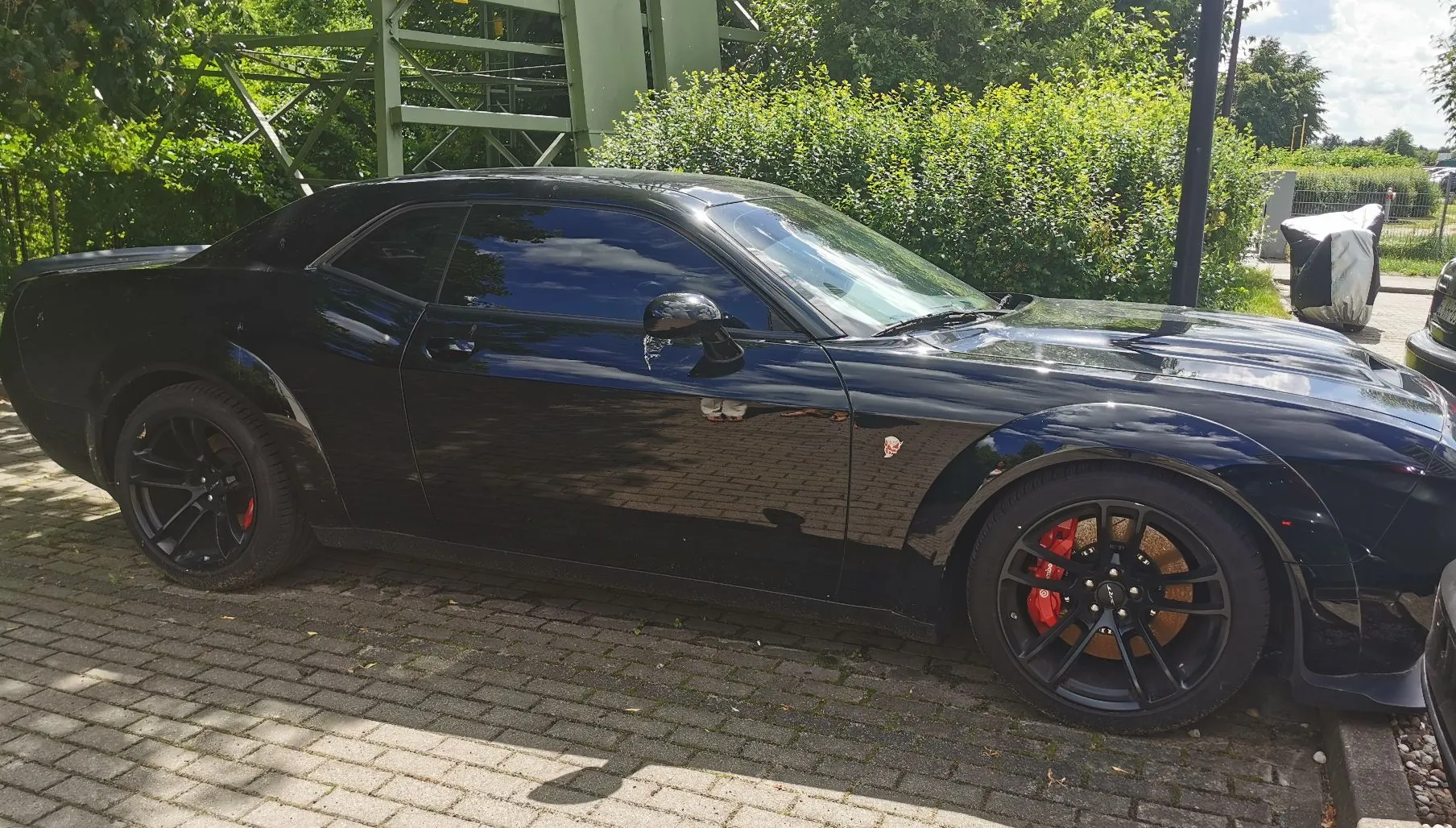 Czarny samochód sportowy Dodge Challenger zaparkowany bokiem na kostce brukowej. Auto posiada czarne felgi z widocznymi czerwonymi zaciskami hamulcowymi. W tle widać zielone krzewy, drzewa oraz fragment zielonej metalowej konstrukcji.