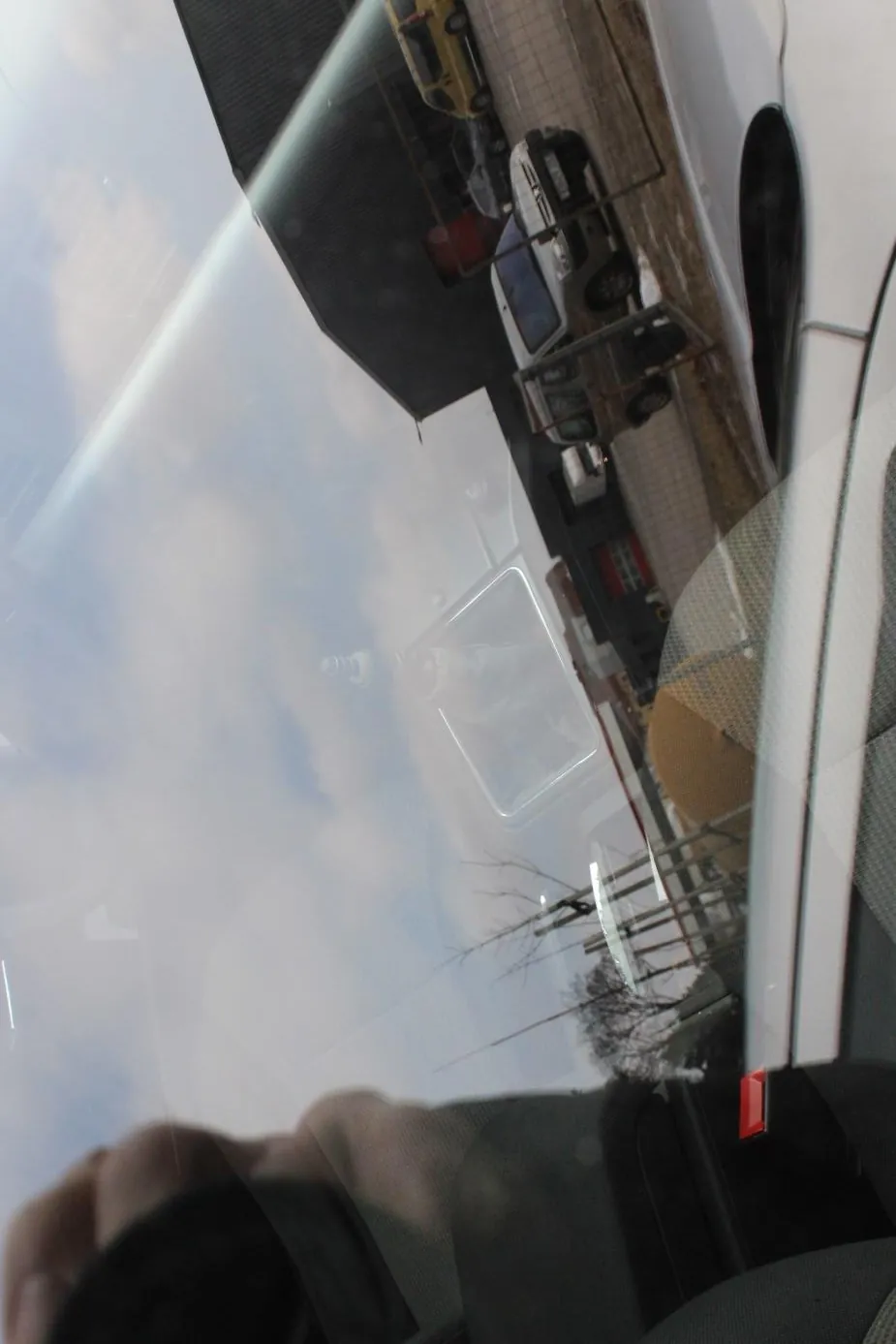 Widok z wnętrza samochodu przez przednią szybę. Po lewej stronie widoczna ręka na kierownicy. Na zewnątrz budynki przemysłowe, zaparkowane pojazdy (biały furgon, żółty samochód), drzewa bez liści, ogrodzenie i częściowo zachmurzone niebo. W odbiciu na szybie fragment wnętrza pojazdu, w tym lusterko wsteczne.