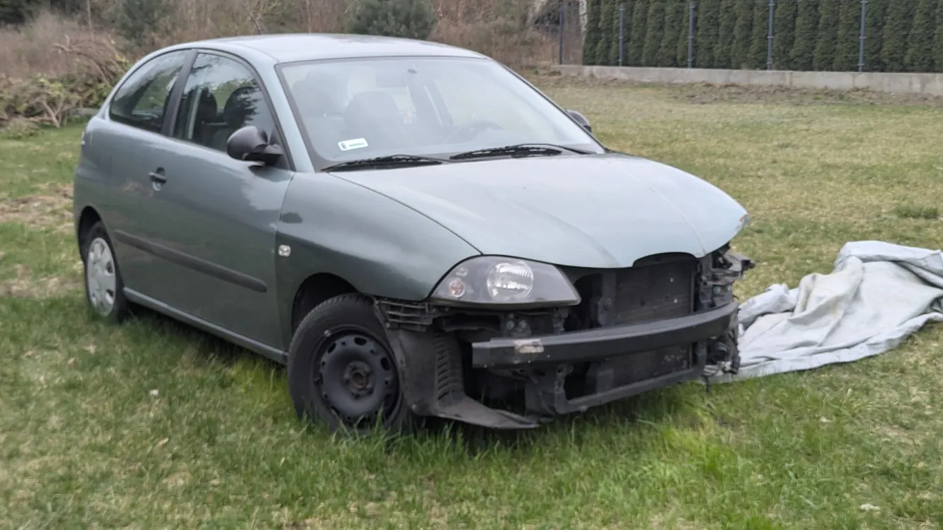 Zniszczony zielony samochód osobowy typu hatchback stojący na trawie. Przednia część pojazdu jest uszkodzona, brakuje zderzaka i części maski. Obok samochodu leży szara płachta materiału. W tle widoczny jest betonowy mur, metalowe ogrodzenie oraz krzewy. Na przedniej szybie znajduje się mała, nieczytelna naklejka.