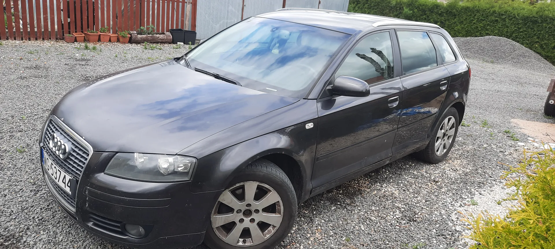Czarny samochód Audi typu kombi zaparkowany na żwirowym podjeździe. Widoczna tablica rejestracyjna MG 53744. W tle czerwone drewniane ogrodzenie z doniczkami z roślinami, metalowa konstrukcja oraz zielone krzewy. Samochód ma srebrne felgi i czyste szyby, na których odbijają się chmury.