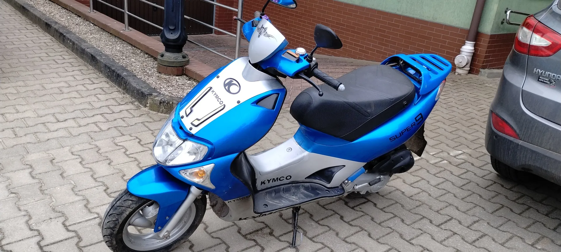 Niebiesko-srebrny skuter marki KYMCO model SUPER 9 zaparkowany na brukowanej powierzchni. Na skuterze widoczne są napisy 'KYMCO' na przedniej części i boku oraz 'SUPER 9' na tylnej części. Po prawej stronie obrazu fragment szarego samochodu marki HYUNDAI z widocznym napisem 'HYUNDAI' na bagażniku. Tło stanowi ceglana ściana budynku z zielonym pasem, metalowe poręcze oraz żwirowa powierzchnia przy krawężniku.