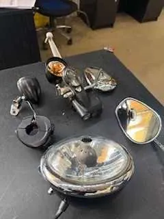 Na czarnym stole ułożone są różne części motocyklowe: okrągły reflektor, kilka lusterków bocznych o różnych kształtach (w tym owalny z odbiciem), czarna rękojeść kierownicy oraz metalowe elementy mocujące. W tle widoczne są fragmenty biurowego krzesła i mebli. Brak czytelnego tekstu.