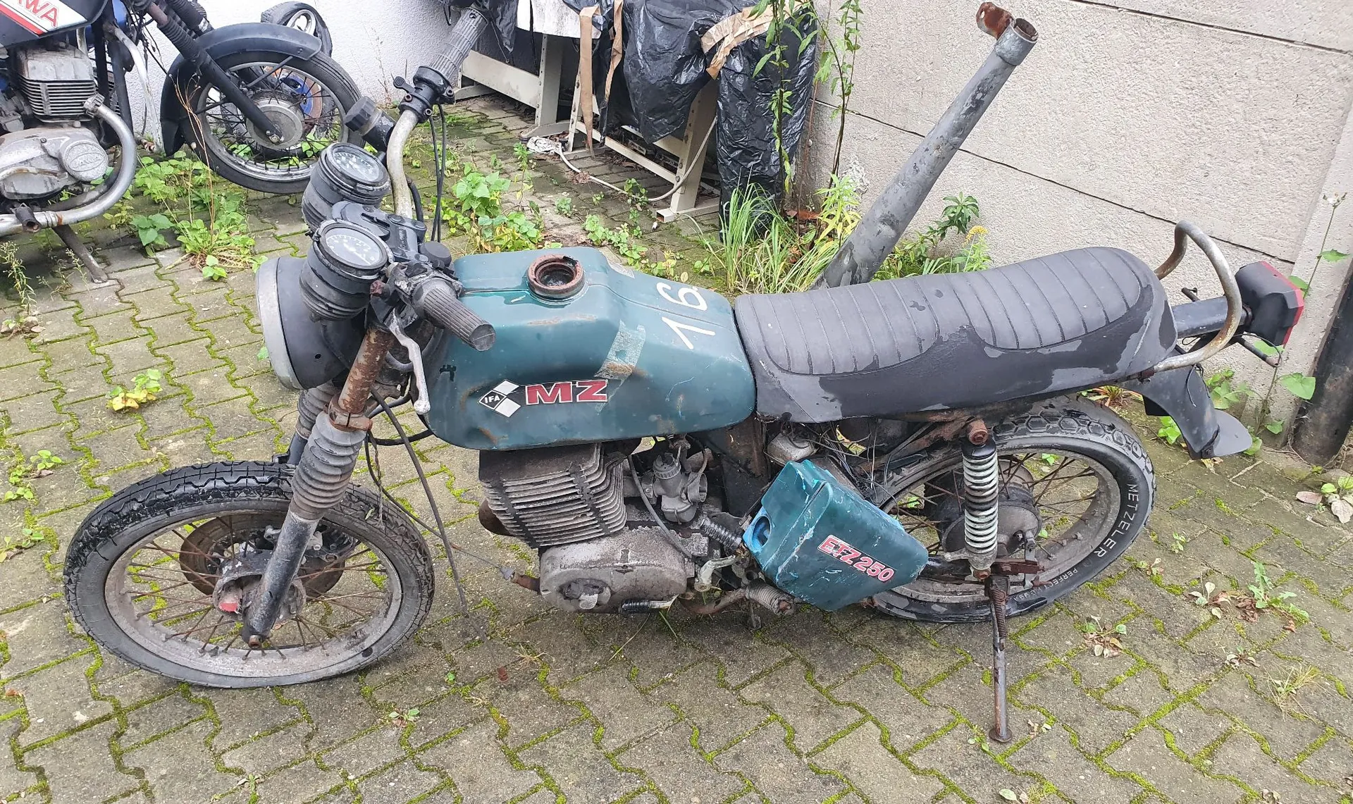 Stary, zużyty motocykl marki MZ w kolorze niebieskim, zaparkowany na kostce brukowej porośniętej mchem. Motocykl ma widoczne ślady rdzy i zabrudzeń. Na baku widnieje napis 'MZ' oraz liczba '76'. Na bocznej osłonie silnika znajduje się napis 'MZ 250'. W tle widoczny jest fragment innego motocykla oraz ściana budynku z workami na śmieci.