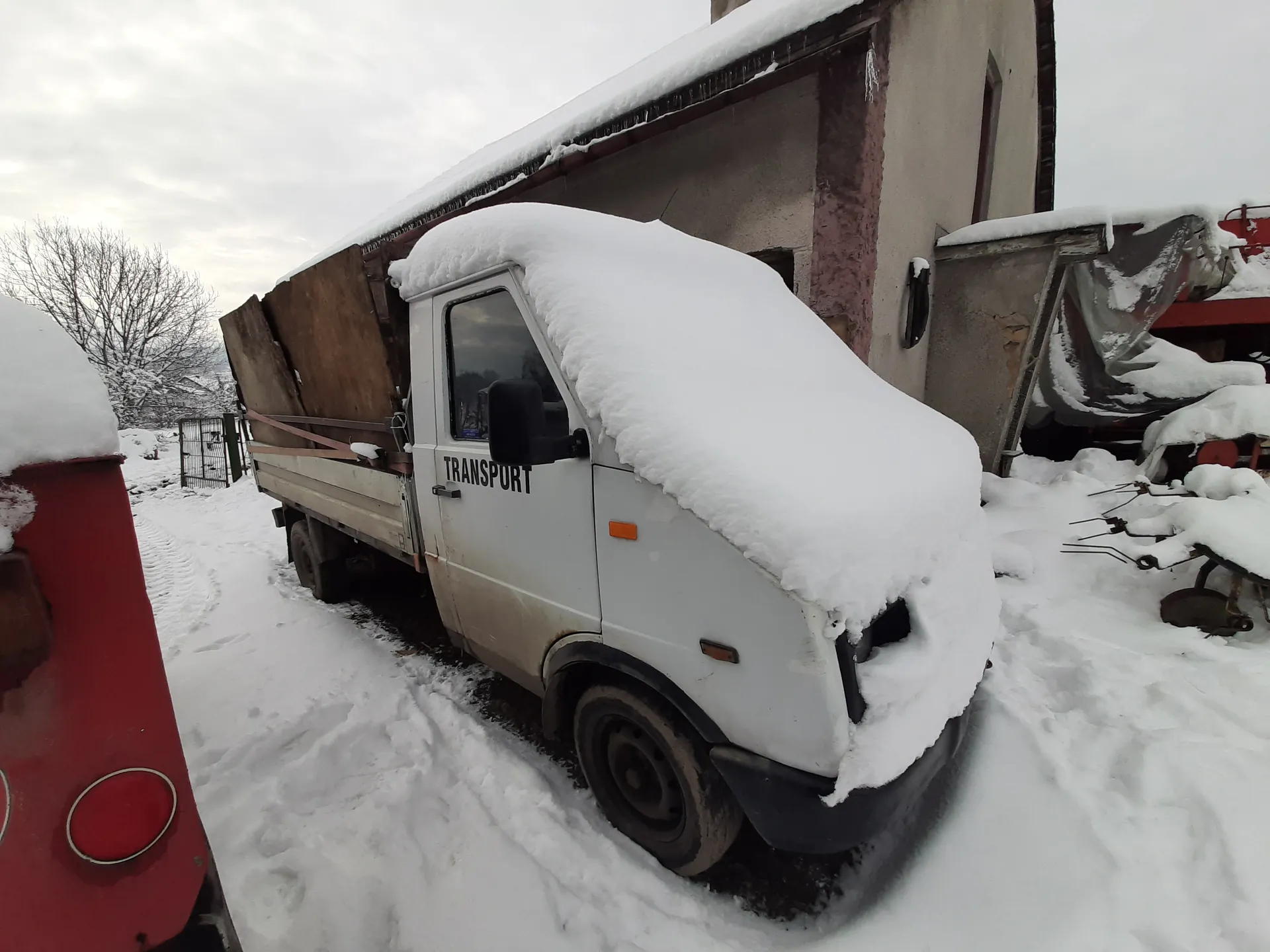 Biały samochód dostawczy z napisem TRANSPORT na drzwiach, pokryty grubą warstwą śniegu. Pojazd stoi na śnieżnej nawierzchni obok budynku o szarej elewacji z czerwoną krawędzią dachu. W tle widoczne są nagie drzewa, fragment czerwonego pojazdu po lewej stronie oraz zakryte śniegiem narzędzia rolnicze po prawej. Niebo jest zachmurzone, a cała scena pokryta śniegiem.