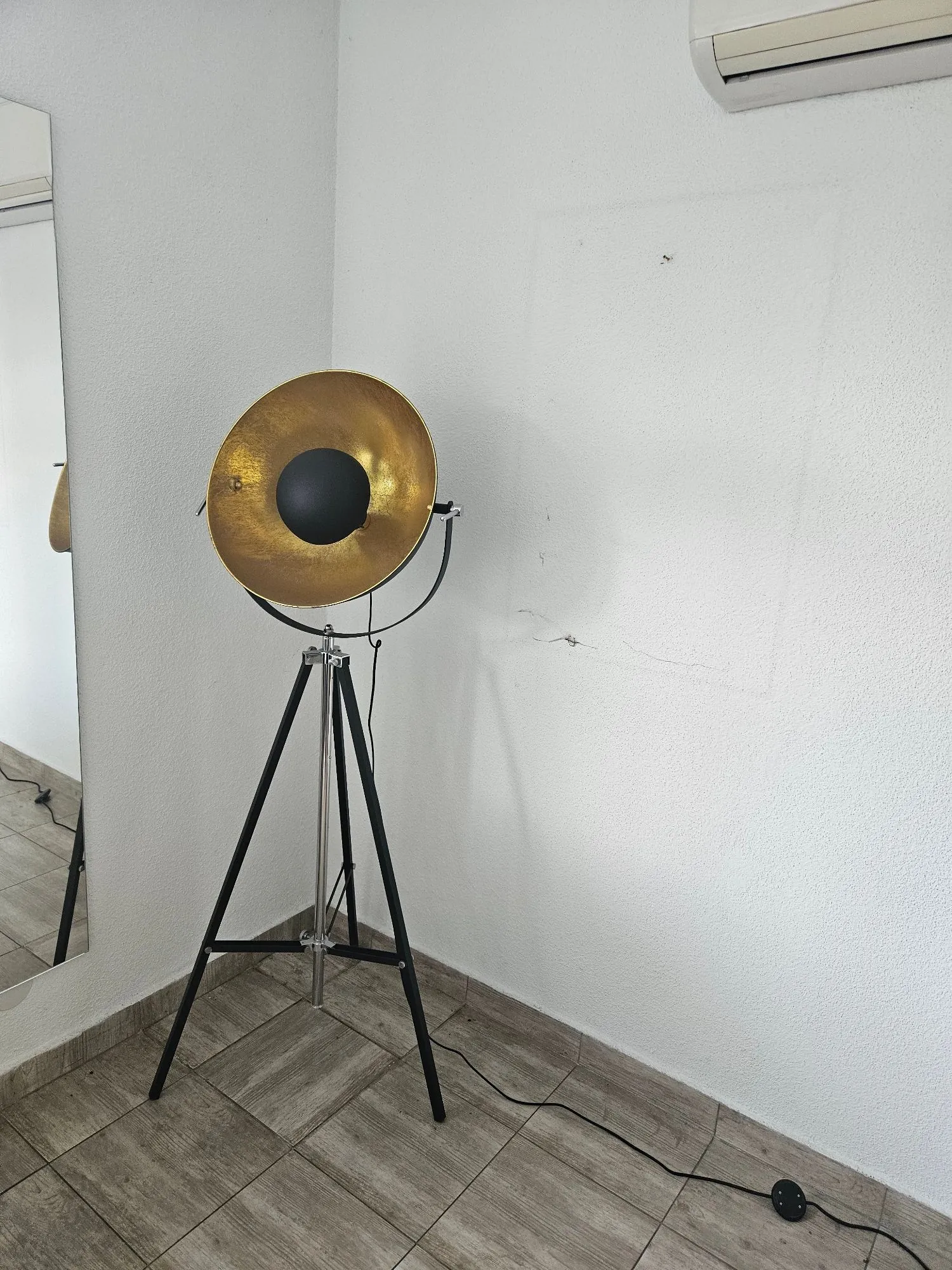 Nowoczesna lampa podłogowa stojąca na czarnym trójnogu. Lampa posiada duży, okrągły klosz w kolorze złotym z czarnym elementem w centrum. Lampa stoi na szarej, kafelkowej podłodze przy białej ścianie. Po lewej stronie widoczne jest lustro, w którym odbija się fragment lampy.