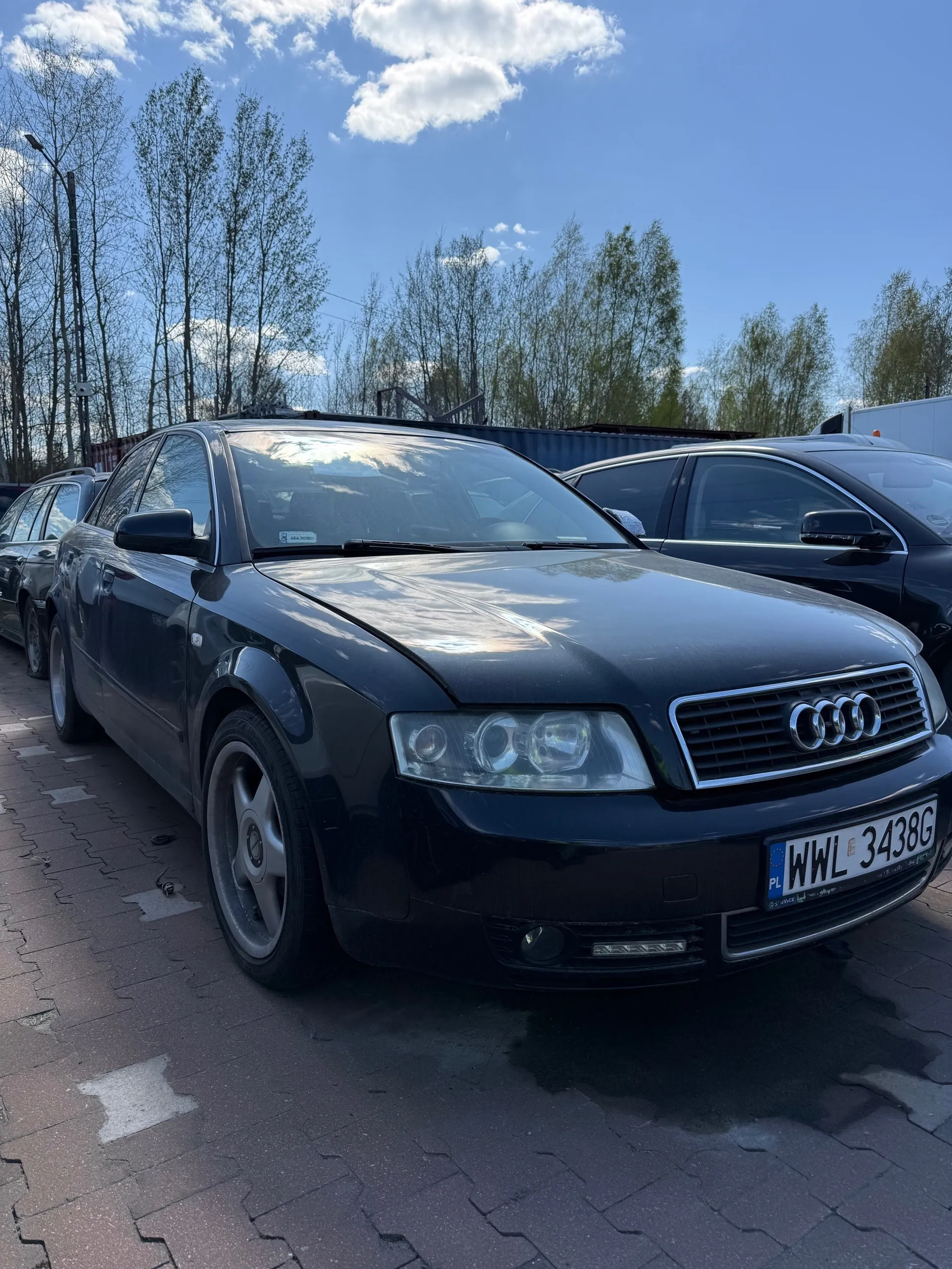 Czarny samochód marki Audi zaparkowany na brukowanej powierzchni. Widoczna tablica rejestracyjna PL WWL 3438G. W tle inne pojazdy, drzewa bez liści, niebo z białymi chmurami oraz kontenery. Samochód ma cztery koła, reflektory i charakterystyczną atrapę chłodnicy z logo Audi.