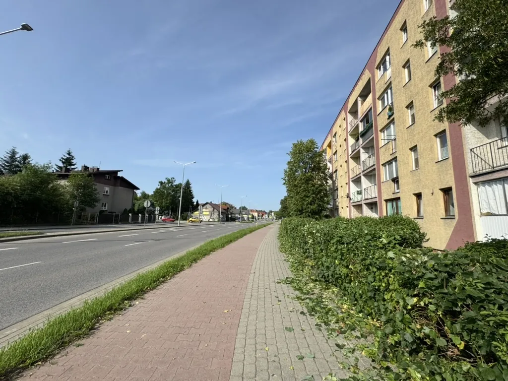 Ulica z chodnikiem wyłożonym czerwonymi i szarymi płytkami, po prawej stronie wysoki blok mieszkalny o żółtej elewacji z czerwonymi akcentami i balkonami, po lewej stronie droga z białymi liniami, drzewa, latarnie uliczne oraz mniejsze budynki w oddali. Na niebie nie ma chmur, widoczne są pojazdy na drodze w oddali.