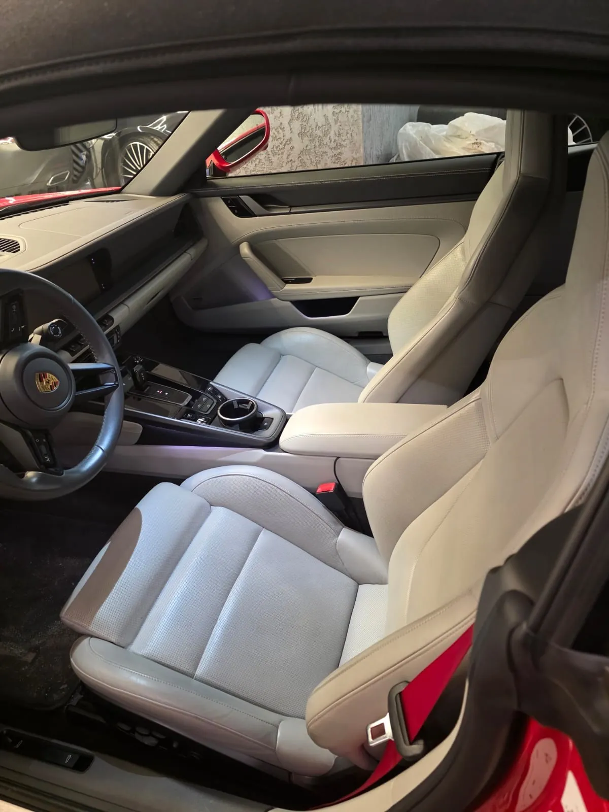 Wnętrze samochodu marki Porsche z jasnymi, beżowymi fotelami. Widoczna kierownica z logo Porsche, deska rozdzielcza, centralna konsola z przyciskami i dźwignią zmiany biegów. Czerwony pas bezpieczeństwa na fotelu pasażera. Przez otwarte drzwi widoczna czerwona karoseria oraz fragmenty innych pojazdów i budynku na zewnątrz.