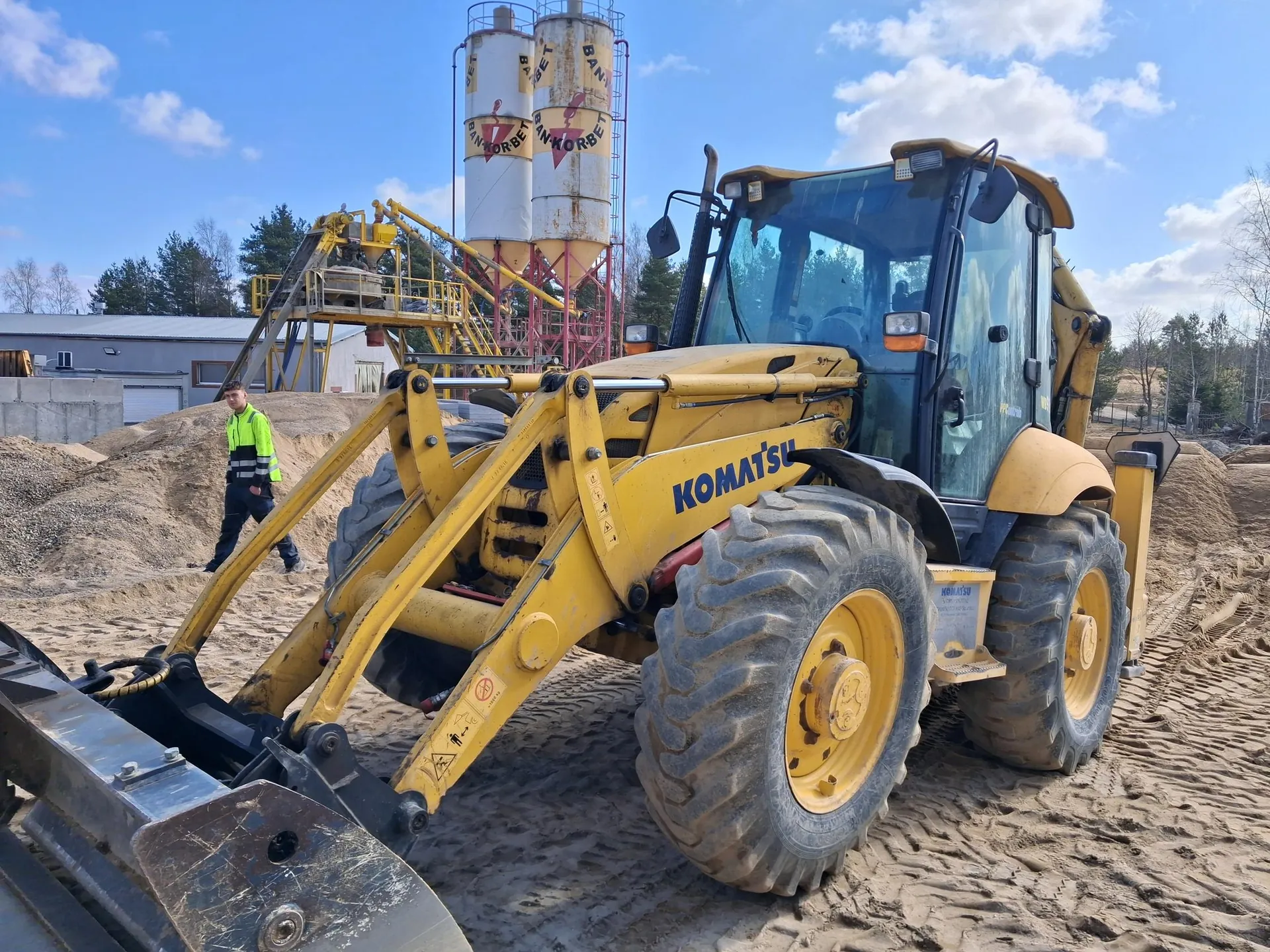 Żółty ładowarka kołowa marki KOMATSU na terenie budowlanym z piaskiem i żwirem. W tle widoczne są dwa wysokie zbiorniki z napisem BANKORBET, przemysłowe konstrukcje, budynek, drzewa oraz niebo z chmurami. Przy maszynie stoi osoba w zielonej kamizelce odblaskowej i ciemnych spodniach. Na maszynie widoczne są duże opony, kierownica i elementy hydrauliczne.