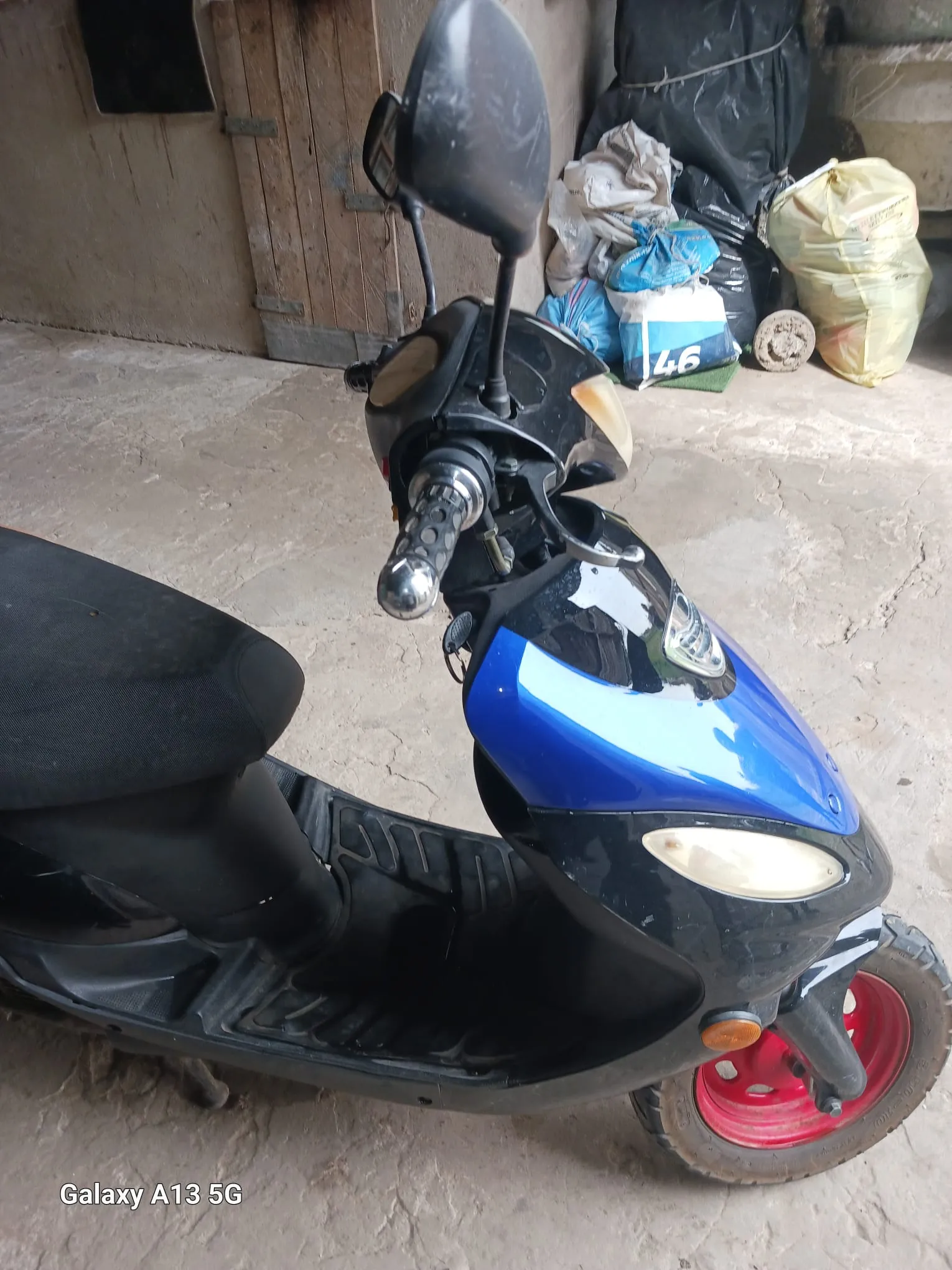 Czarno-niebieski skuter z czerwonymi felgami, widoczne lusterko, kierownica z czarną obudową i reflektorem. W tle drewniane drzwi, worki (w tym niebieski z białą cyfrą 46), żółty worek i betonowa podłoga. W lewym dolnym rogu widoczny napis Galaxy A13 5G.