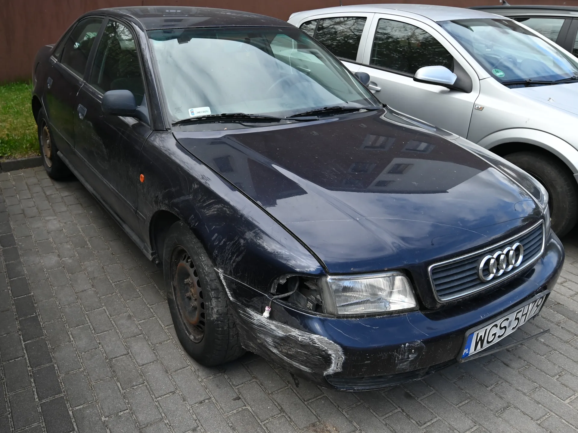 Czarny samochód marki Audi z widocznymi uszkodzeniami na przednim lewym zderzaku i reflektorze. Na masce widoczne ślady zarysówania. Tablica rejestracyjna WGS 5H9 z oznaczeniem PL. Po prawej stronie częściowo widoczny srebrny samochód. Tło: brukowana powierzchnia, trawa i brązowa ściana. Na przedniej szybie czarnego Audi naklejka z nieczytelnym tekstem.