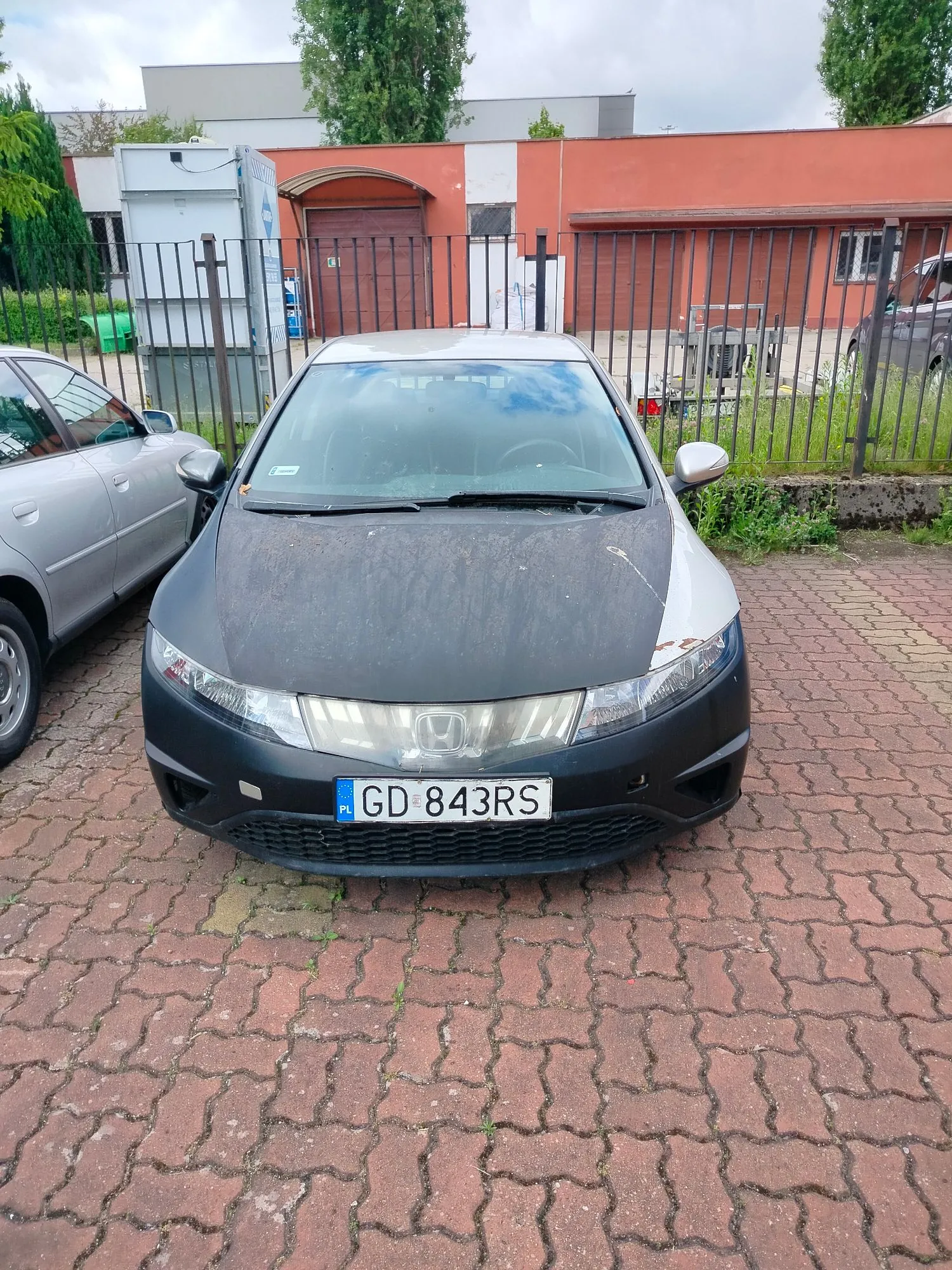 Srebrny samochód marki Honda zaparkowany przodem na kostce brukowej. Maską samochodu pokrywa się ciemna, matowa folia lub farba, która jest częściowo zdrapana po prawej stronie. Samochód ma polską tablicę rejestracyjną o numerze GD 843RS. W tle widoczny jest metalowy płot, czerwony budynek oraz zielone drzewa.