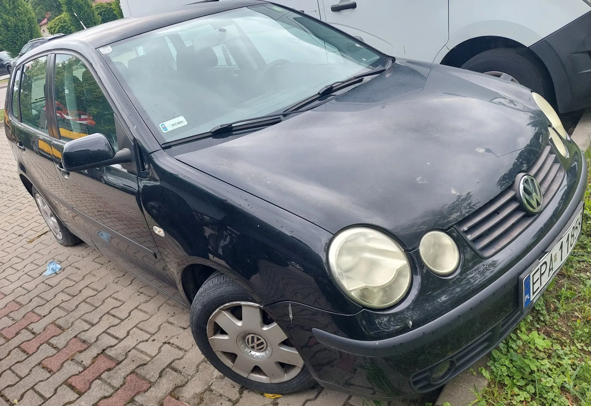 Czarny samochód osobowy marki Volkswagen Golf zaparkowany na kostce brukowej. Pojazd jest widoczny z przodu i z boku, ma widoczne zmatowiałe reflektory oraz tablicę rejestracyjną zaczynającą się od liter EPA. W tle widoczny jest fragment białego samochodu.