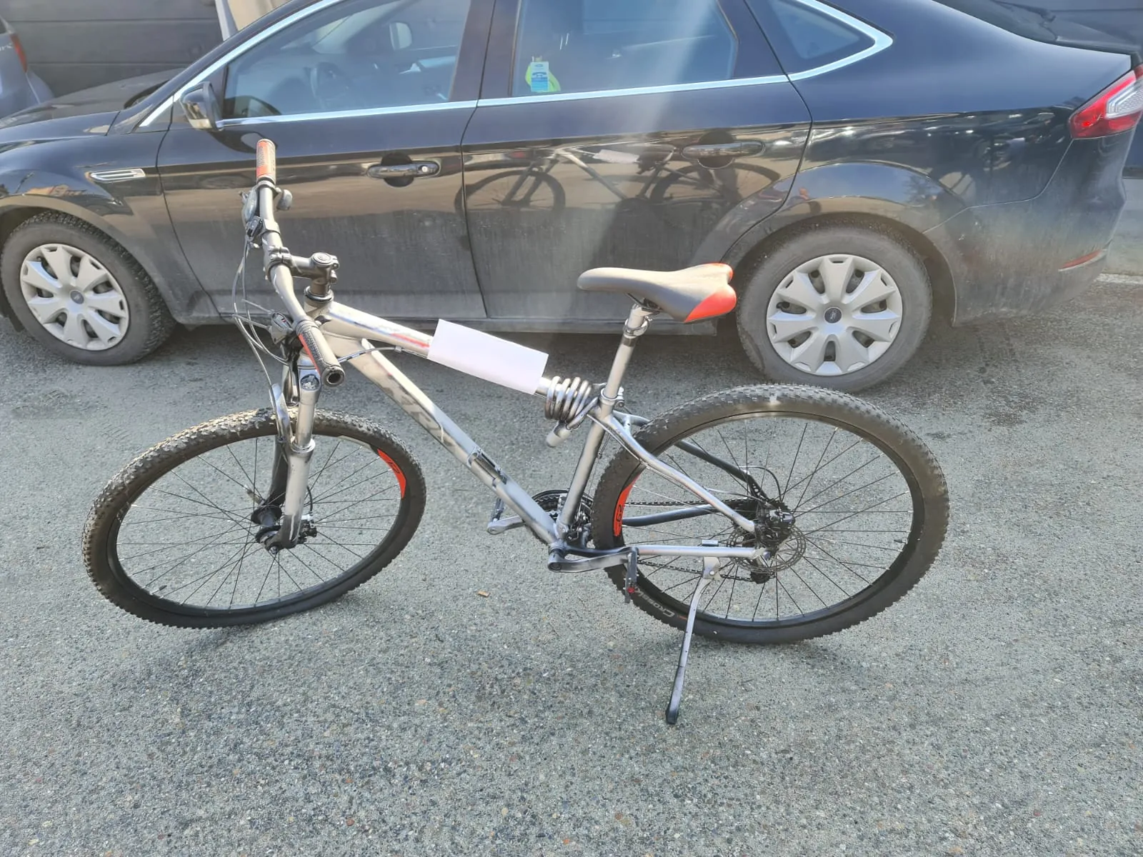 Srebrny rower górski z czerwono-czarnym siedziskiem i białym kartonikiem przy ramie, stojący na podkładowym asfalcie. W tle ciemnoszary samochód osobowy z srebrnymi felgami, zaparkowany na ulicy. Na szybie tylnej samochodu widoczny mały kolorowy naklejka.