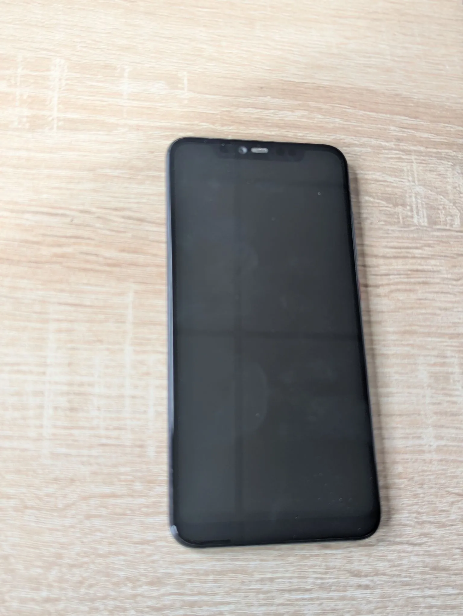Czarny smartfon z wyłączonym ekranem, leżący pionowo na jasnym, drewnianym blacie o widocznym usłojeniu.