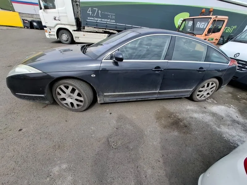 Ciemnoszary samochód osobowy typu sedan zaparkowany na asfaltowej powierzchni. W tle widoczne są inne pojazdy: biała ciężarówka z zielonym przyczepem z napisami '4.7t', 'www.berger-ecotrail.com' oraz pomarańczowy pojazd budowlany z częściowo widocznym napisem 'B RGE rail'. Na pierwszym planie fragment białego samochodu.