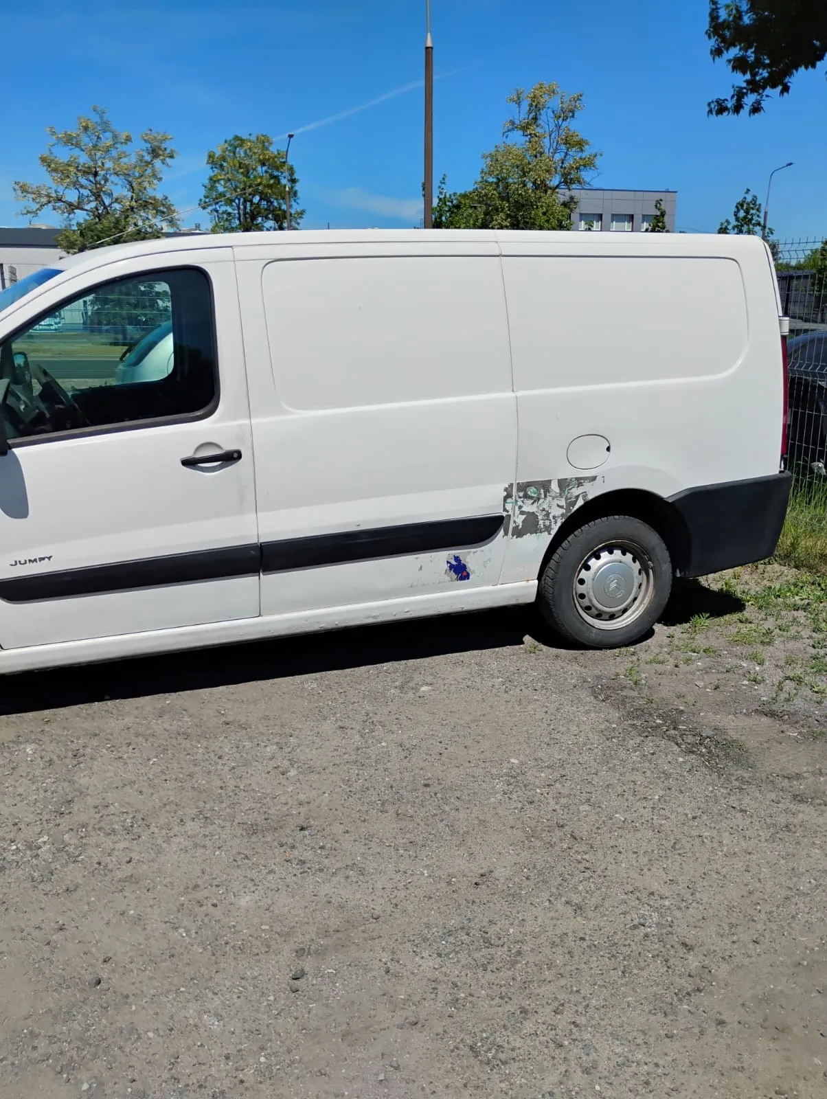 Boczny widok białego vana marki Citroën Jumpy zaparkowanego na żwirowym podłożu. Na dolnej części nadwozia, nad tylnym kołem, widoczne są uszkodzenia lakieru i odpryski. W tle widać niebieskie niebo, zielone drzewa oraz fragmenty budynków.